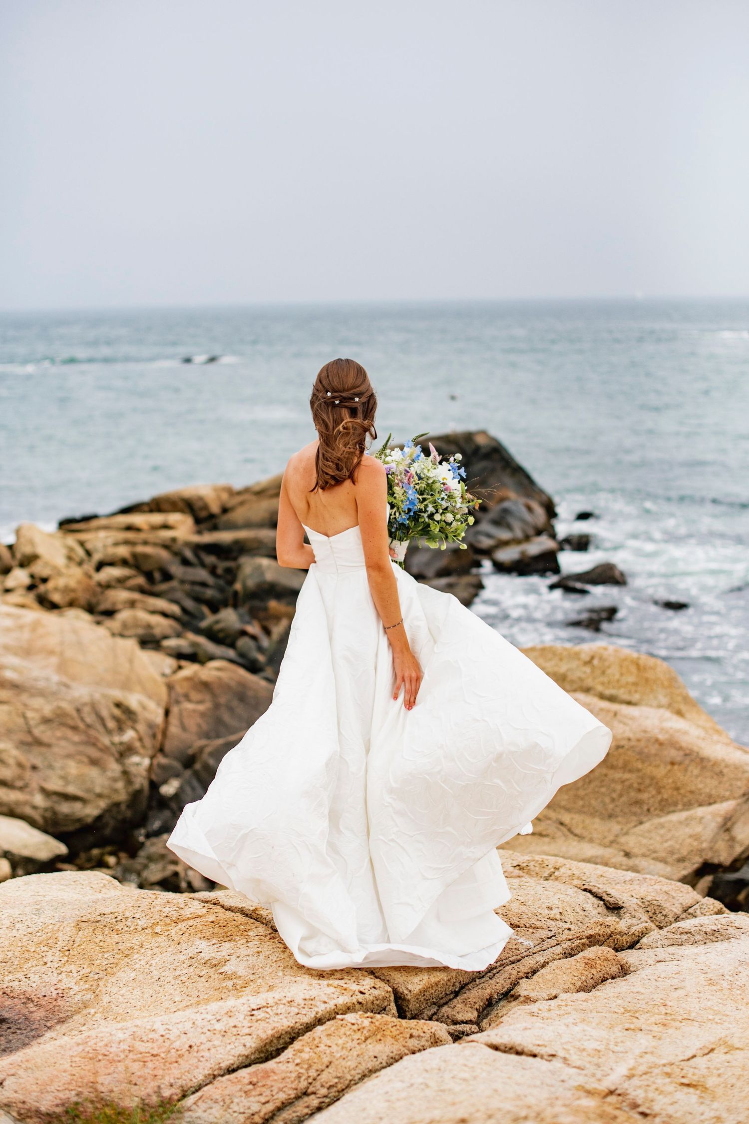 A bride in a flowing white dress walks along coastal rocks by the ocean.