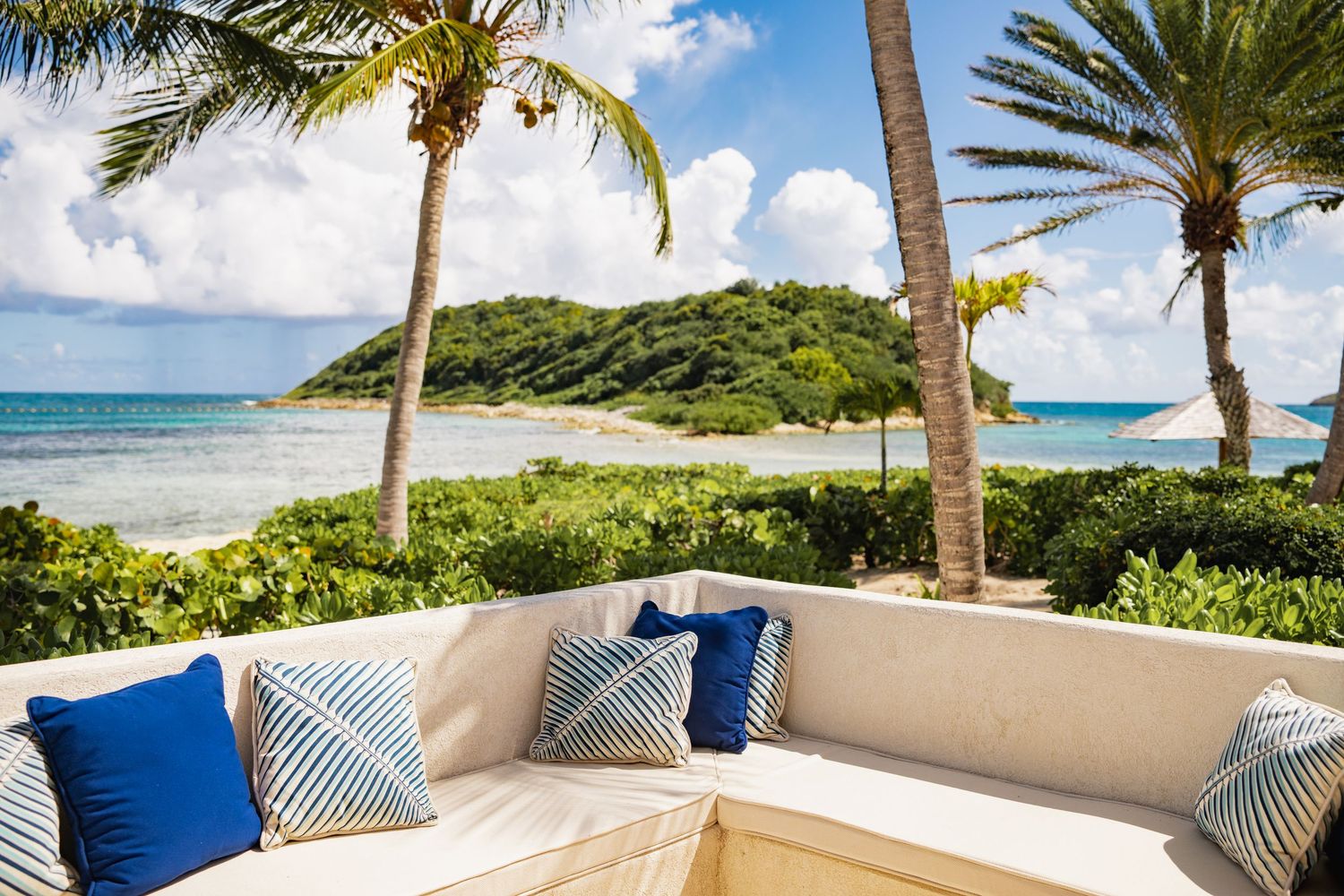 Luxurious outdoor seating area with blue cushions overlooking a tropical beach with palm trees and turquoise waters.