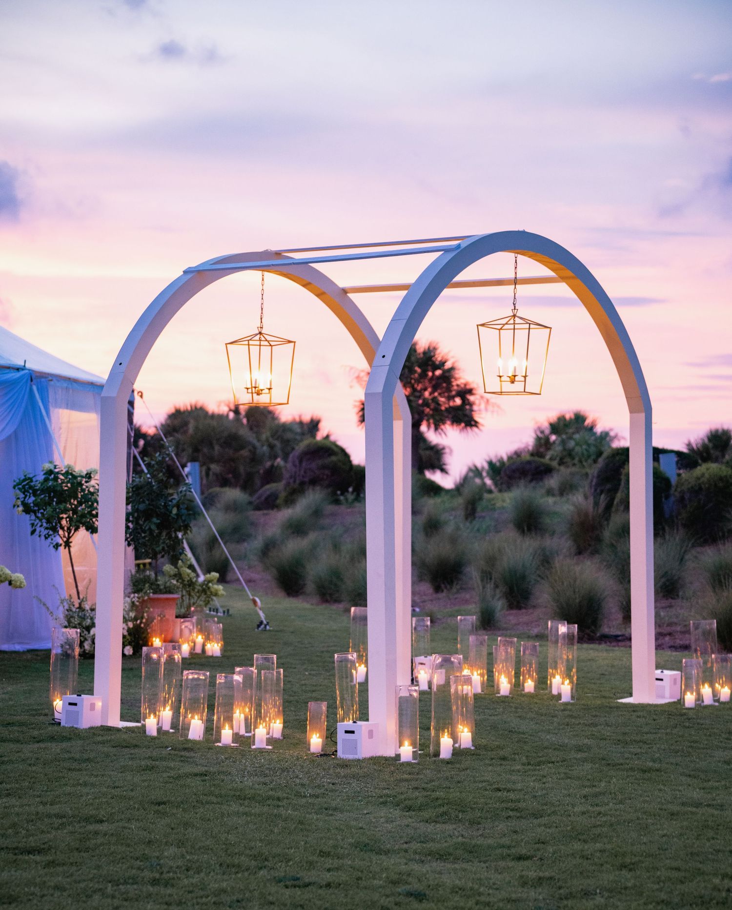 White arched wedding ceremony backdrop with hanging lanterns and candles at sunset.