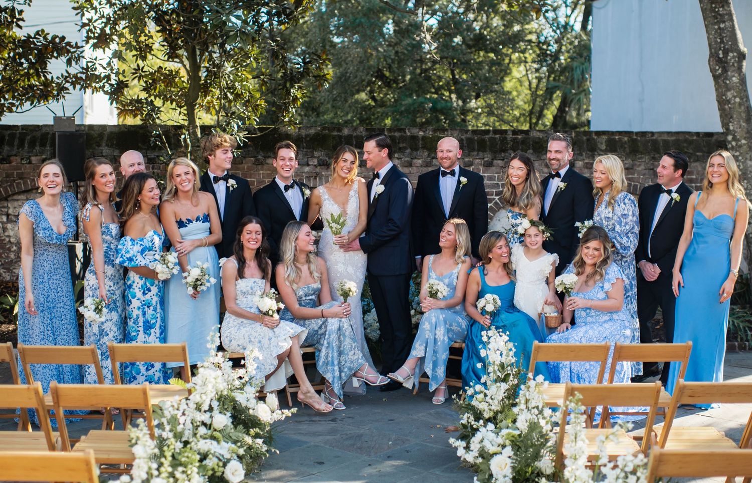 Wedding party in light blue bridesmaid dresses and dark suits pose together outdoors on a sunny day.