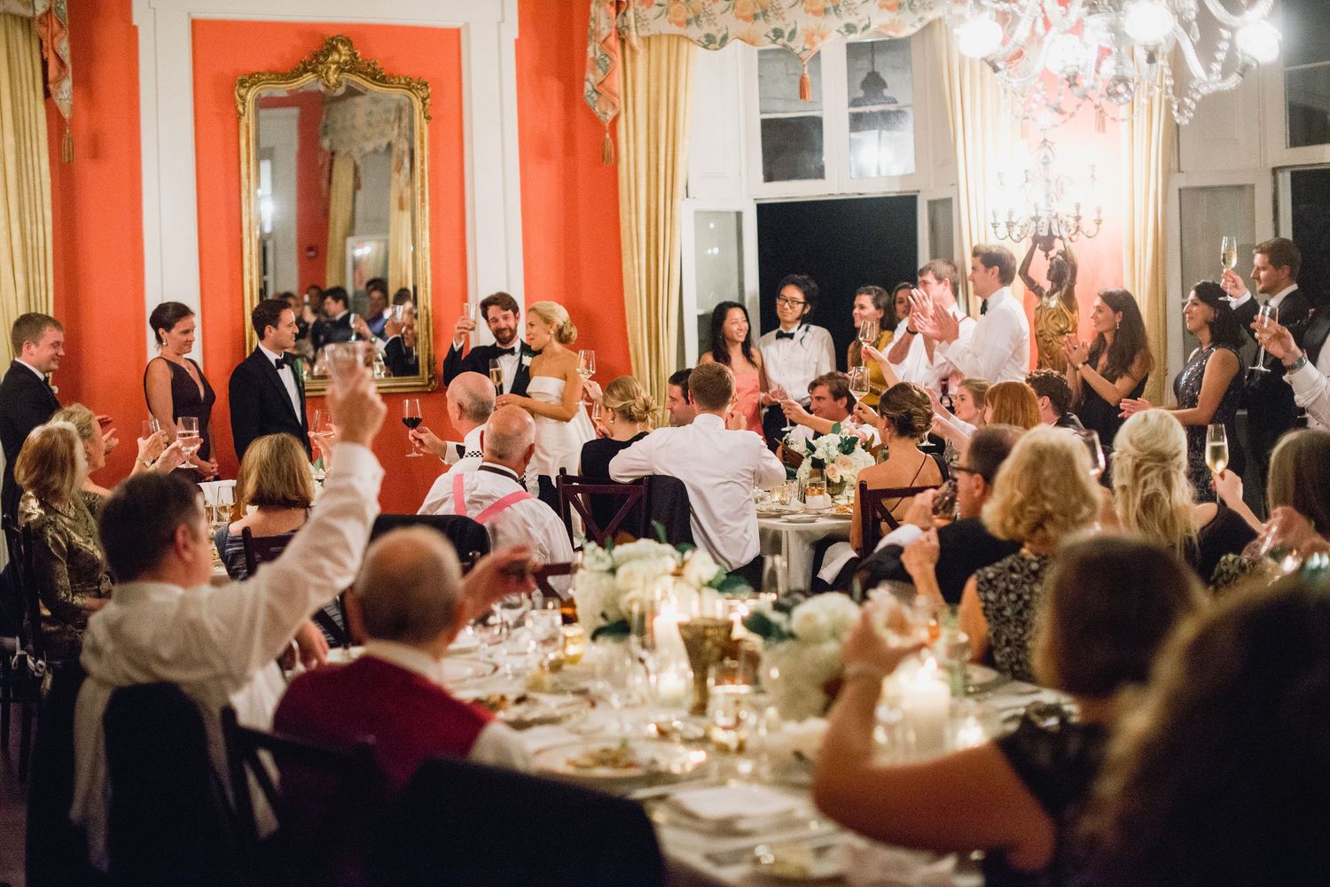 Elegant wedding reception in a grand ballroom with orange walls and crystal chandeliers as guests dine at decorated tables.