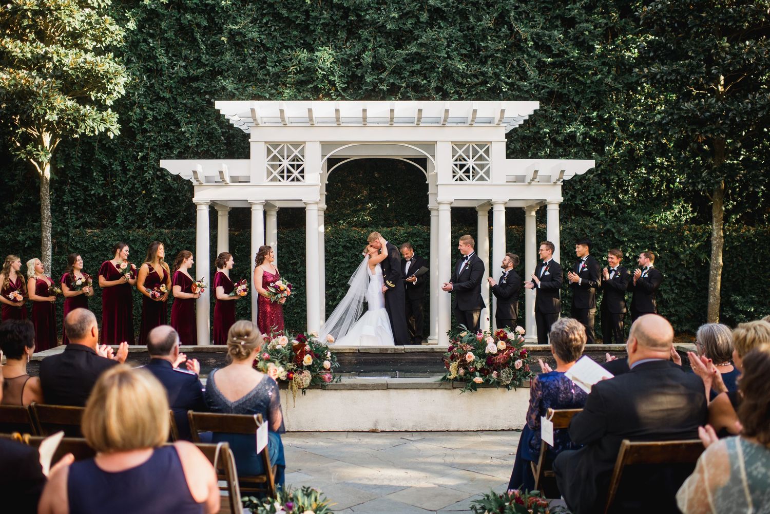 A wedding ceremony takes place outdoors under a white pergola with guests seated on wooden benches in a garden setting.