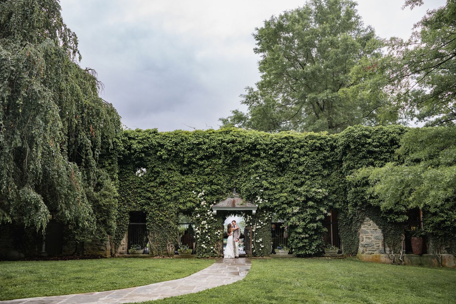 A winding garden path leads through lush greenery and hedges under an overcast sky.