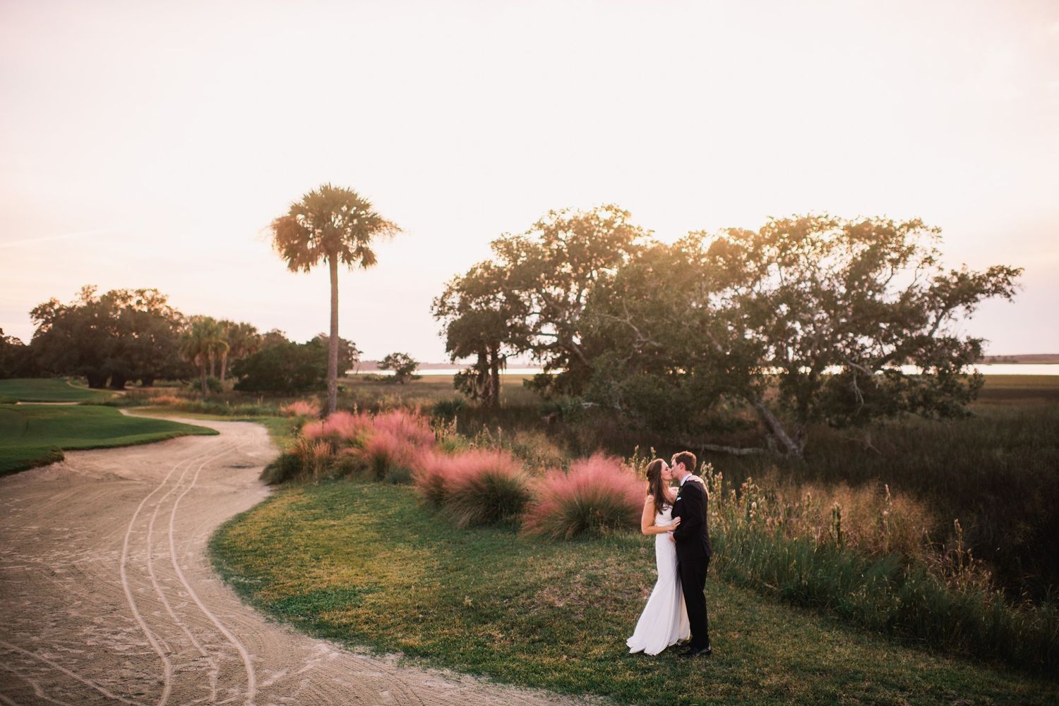 A couple stands together on a winding path at sunset with tall grasses and trees in the background.