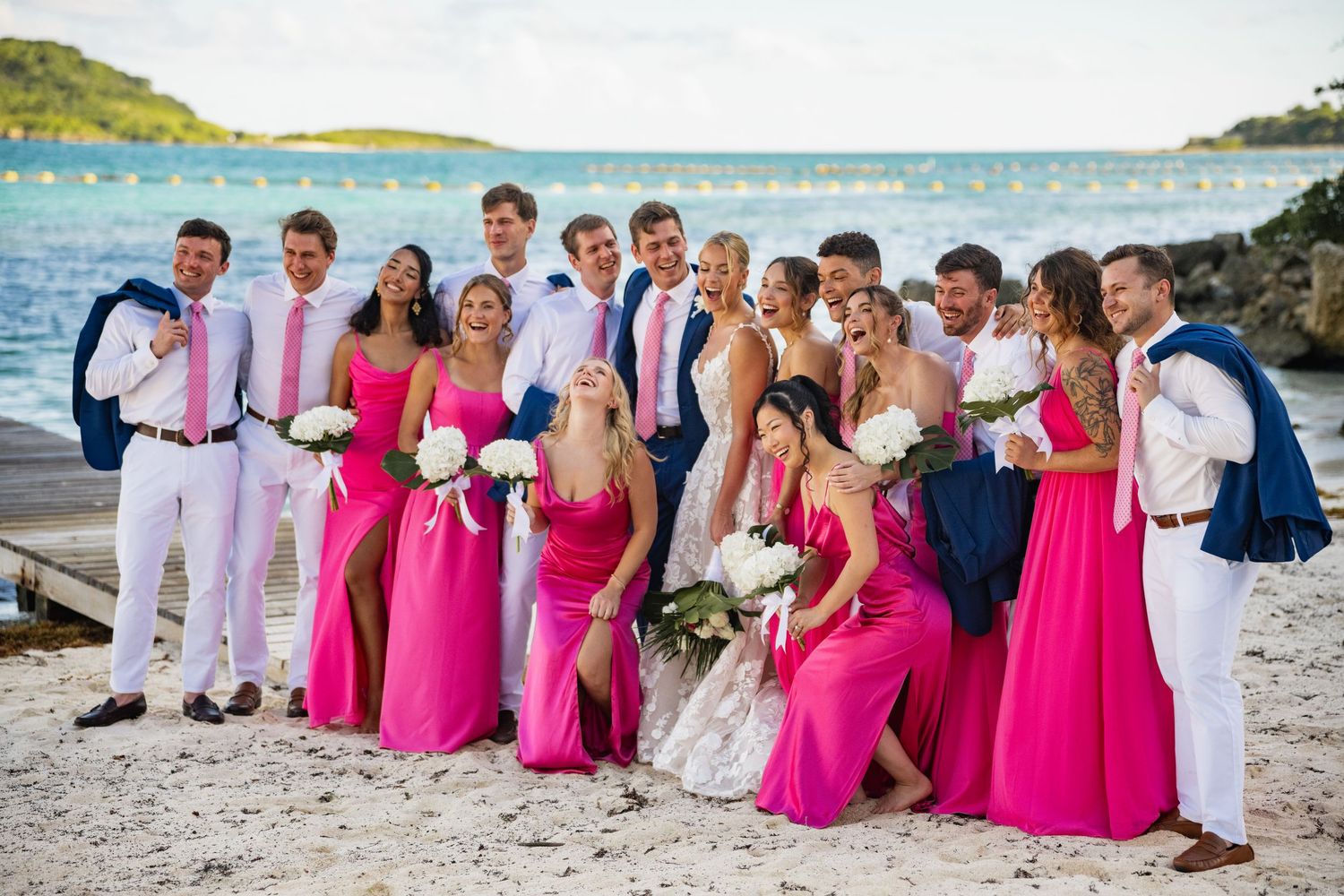 Wedding party in pink bridesmaid dresses and white attire posing together on a tropical beach.