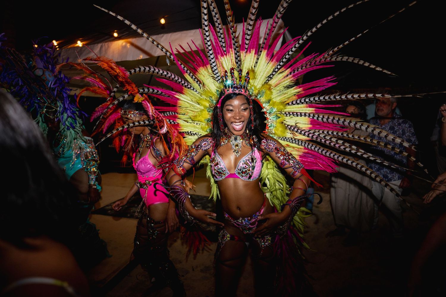 Colorful carnival dancers perform in vibrant feathered costumes with bright pink, yellow and blue plumes on stage.