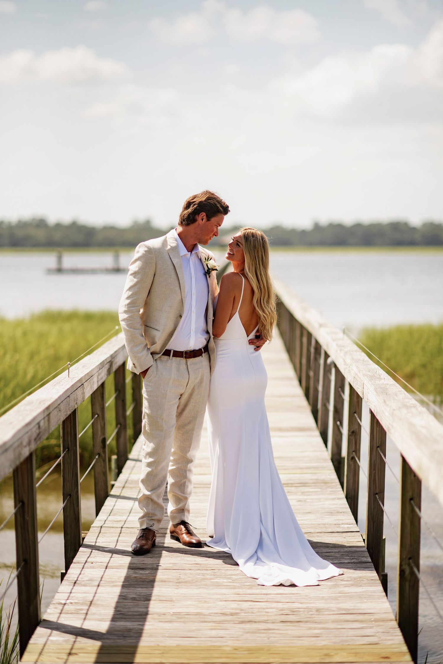 A couple in white attire share romantic moments on a wooden boardwalk overlooking a scenic waterfront.