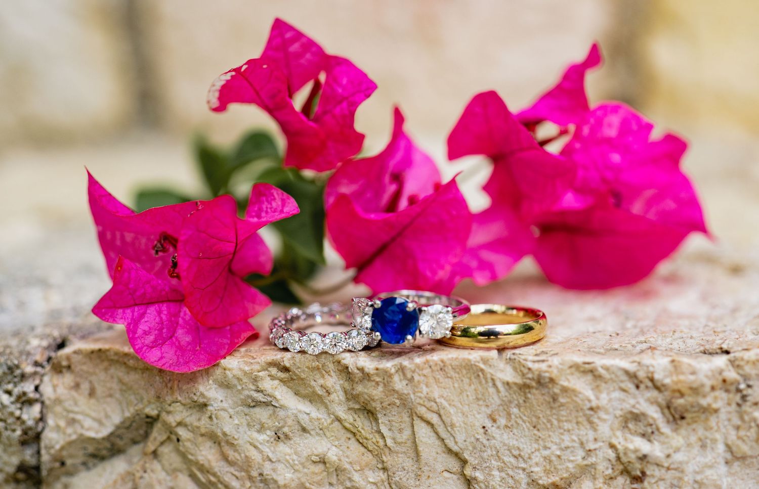 Wedding rings with sapphire and diamonds displayed on stone with vibrant pink bougainvillea flowers.
