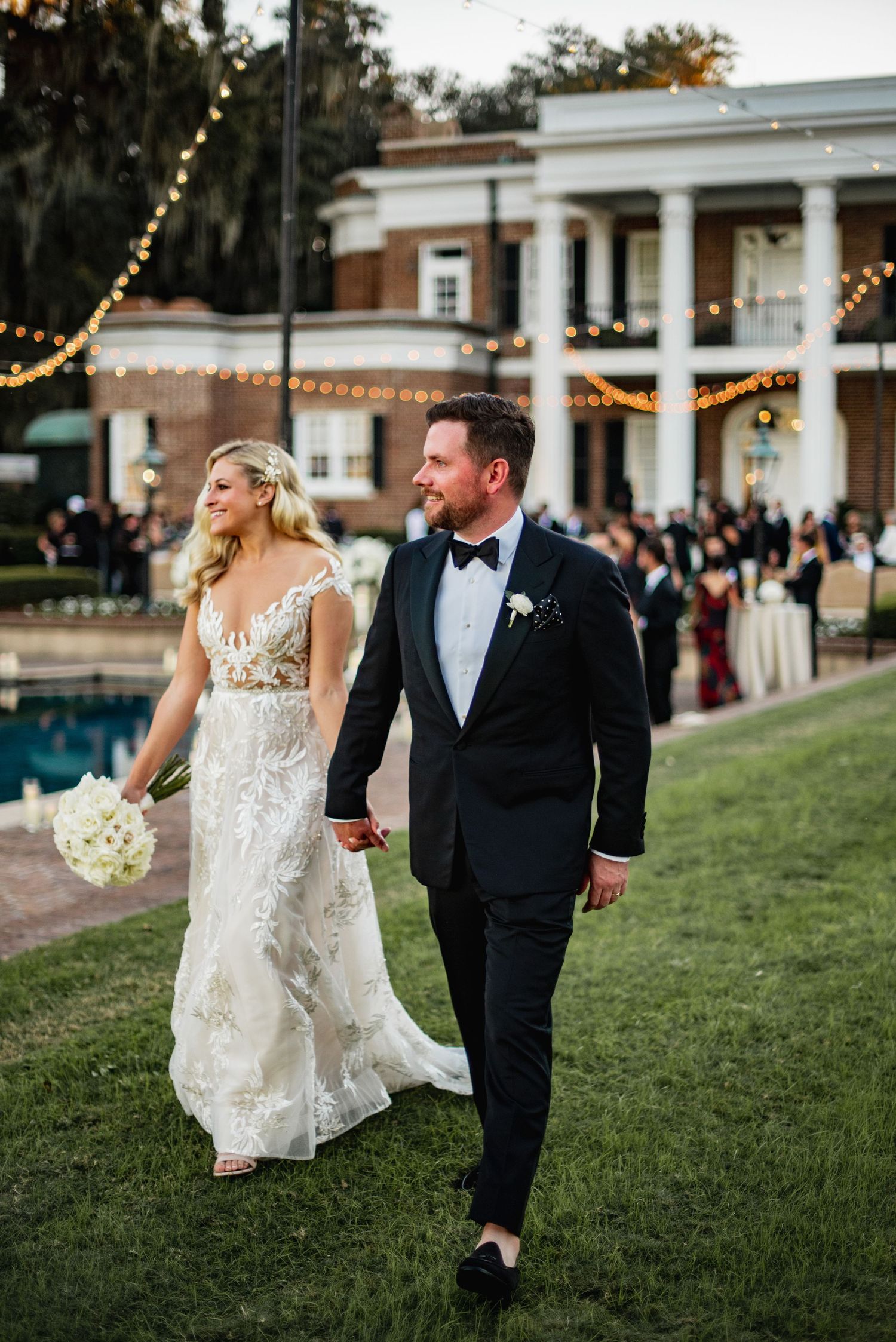 A couple walks together on a grassy lawn outside a classic columned building with string lights at dusk.