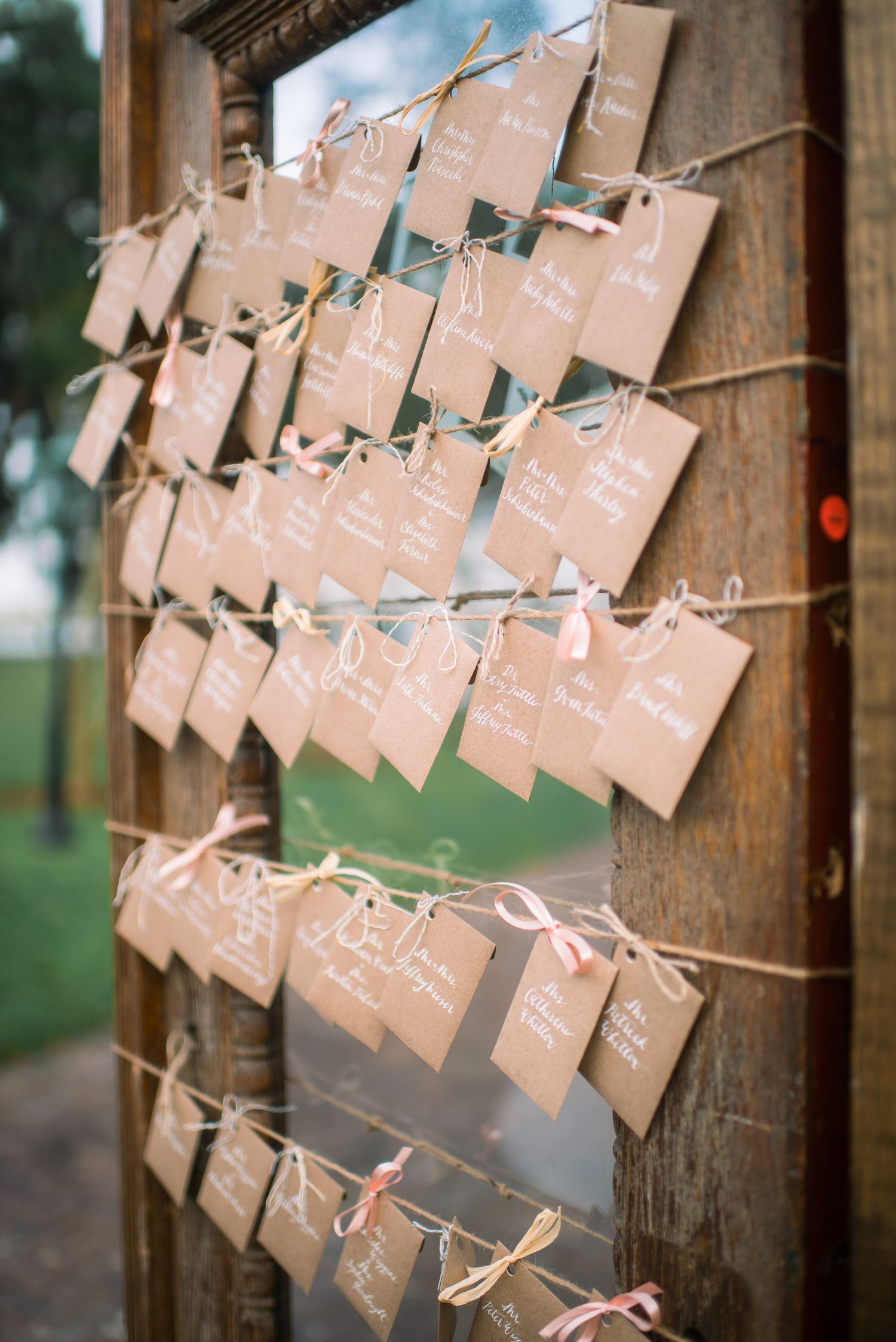 Rustic wedding escort cards with names written in calligraphy hang from twine against a wooden post in soft lighting.