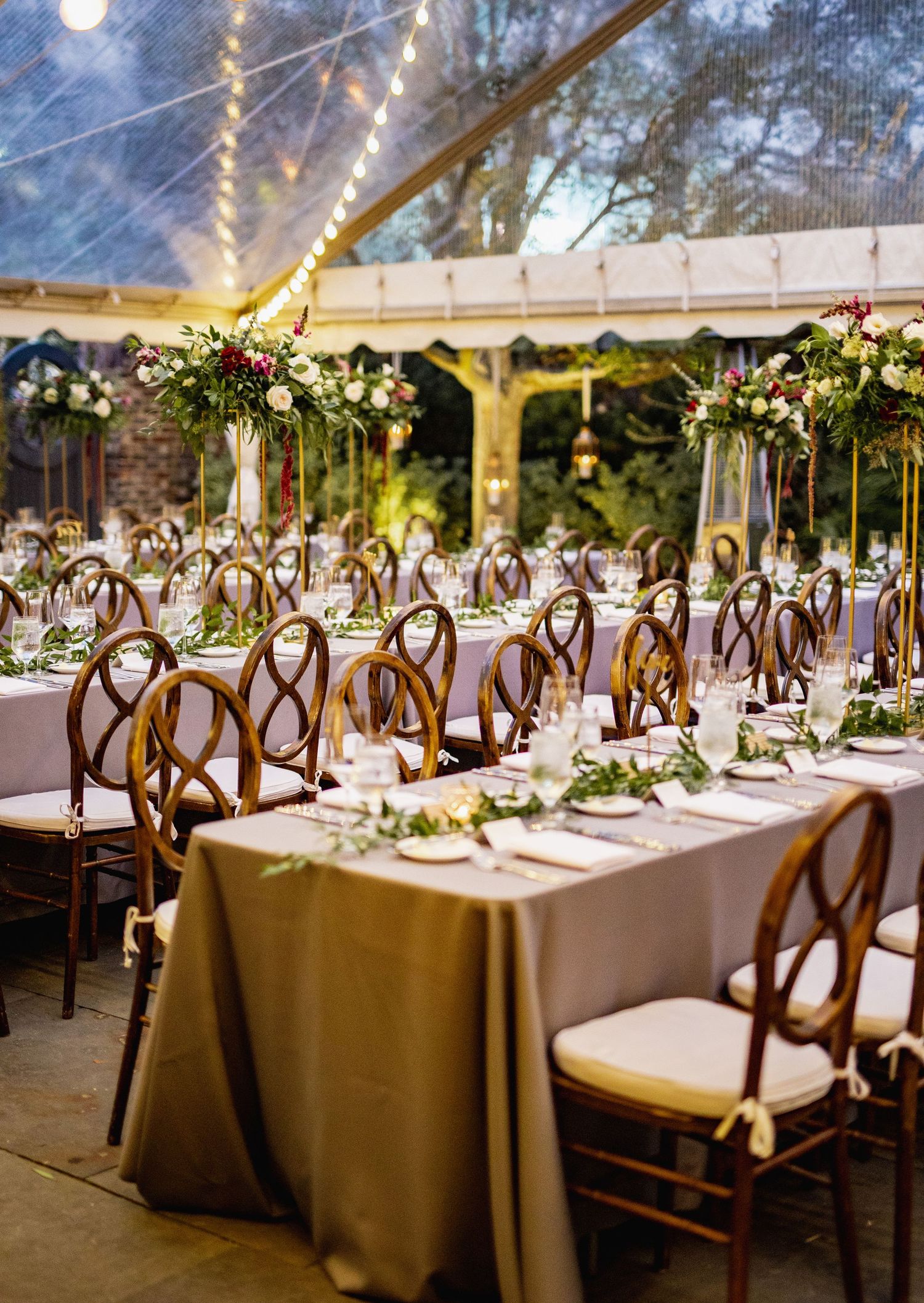Elegant wedding reception tables set with wooden cross-back chairs under a clear tent with twinkling lights and floral centerpieces.