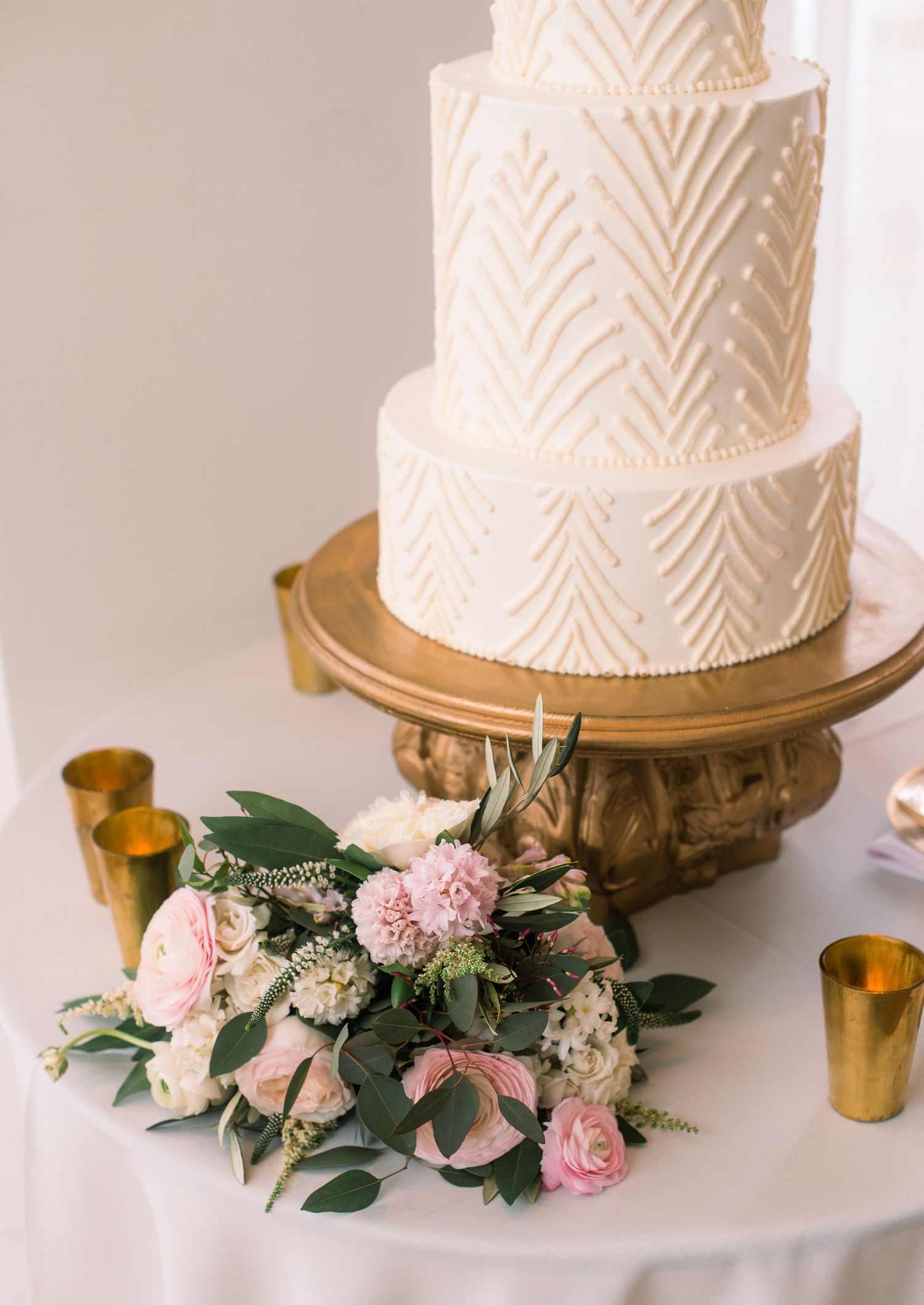 Elegant white wedding cake with chevron pattern and pink floral decorations displayed on a gold cake stand.