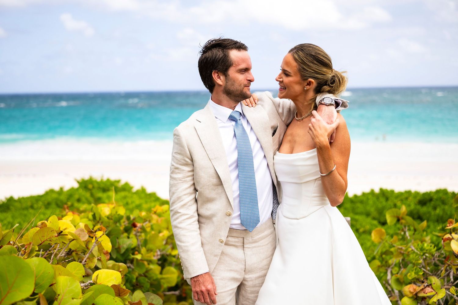 Beachside wedding couple in white attire against turquoise ocean and tropical foliage.