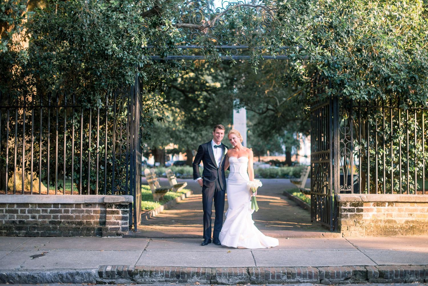 A couple in wedding attire poses together in a romantic garden setting with wrought iron gates and brick walls.