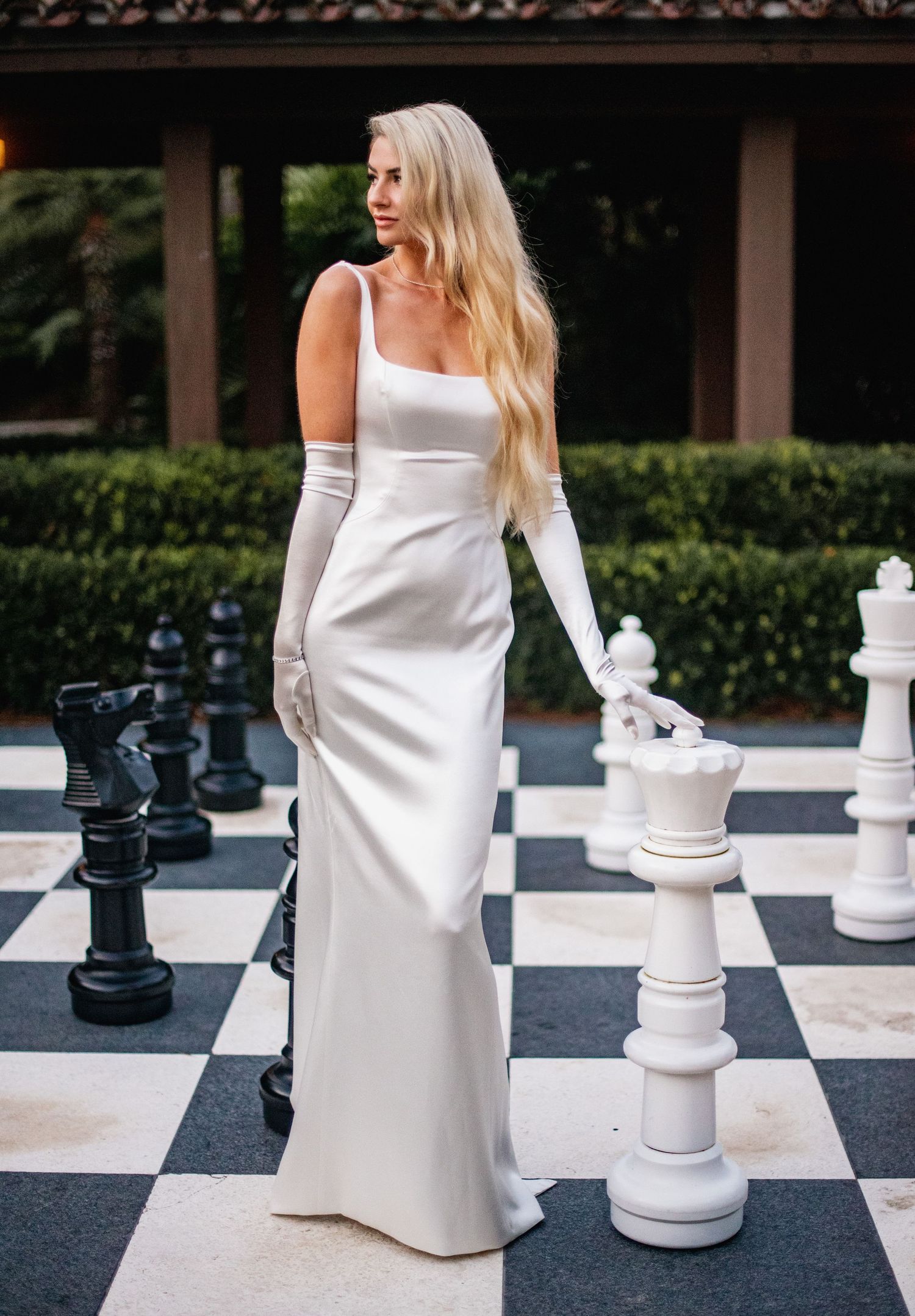 A person in a white dress poses on a life-size outdoor chess board with large black and white chess pieces.