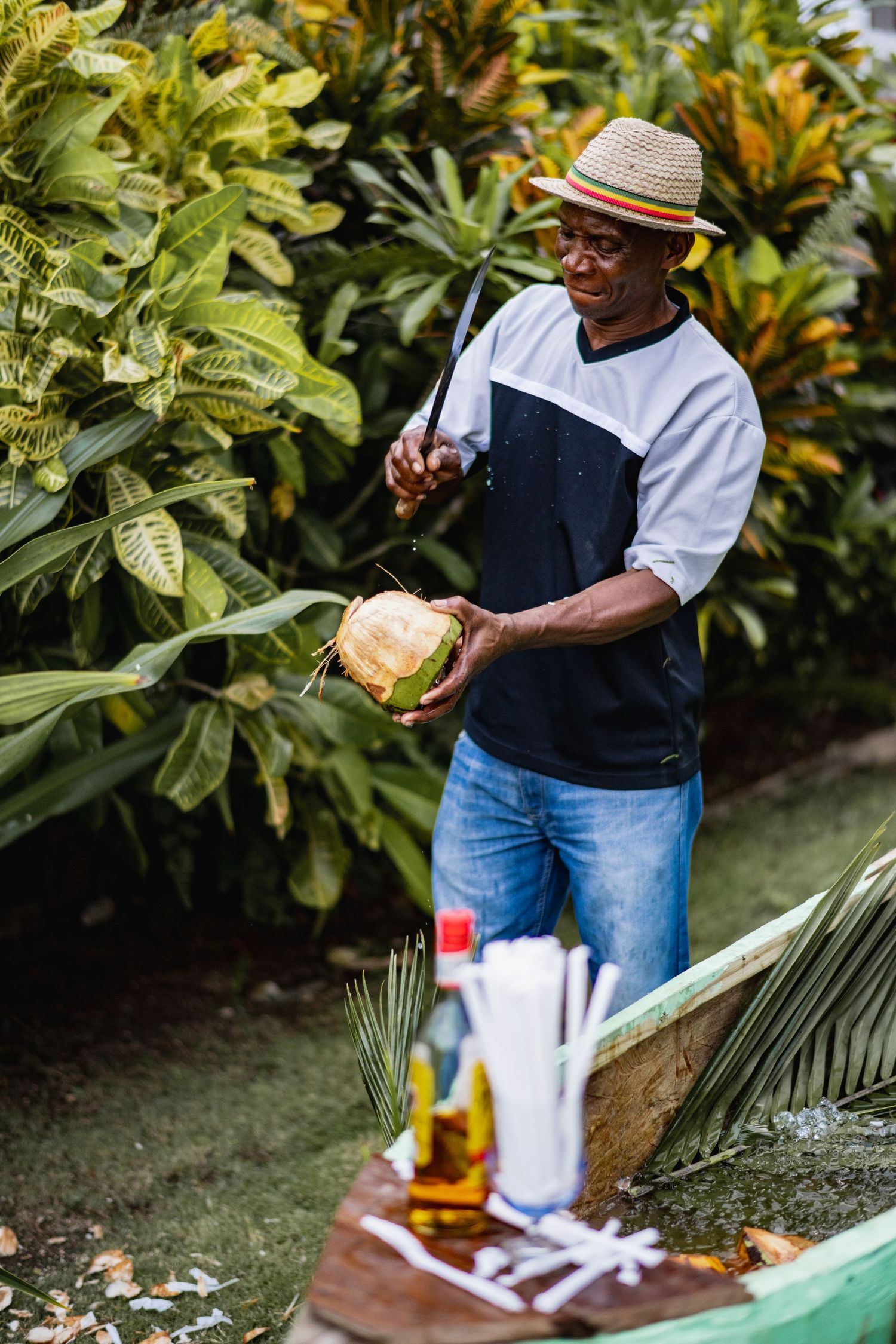 Person in casual attire and straw hat enjoying drinks and conversation in a lush garden setting.