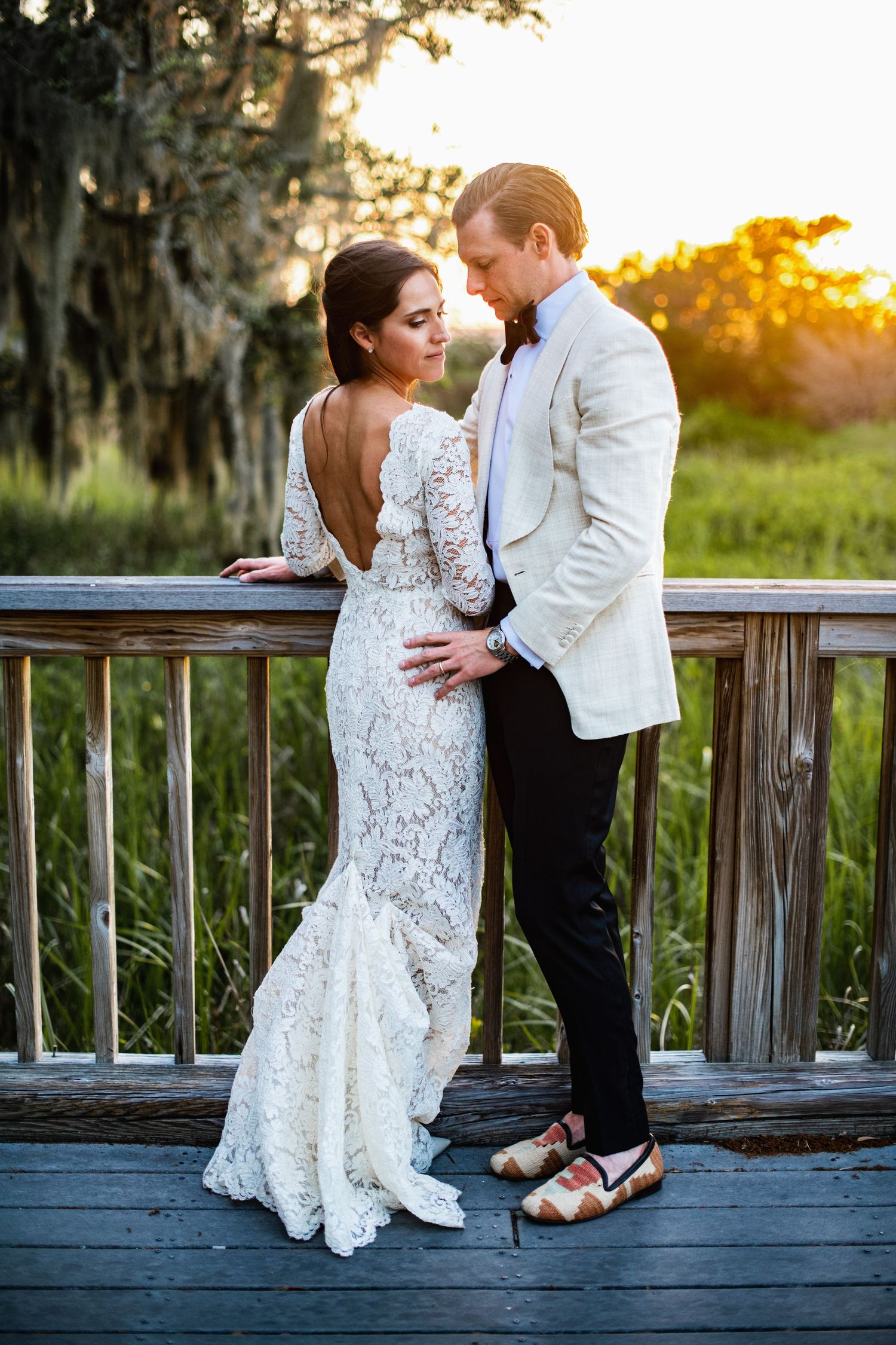 Newlyweds in white lace gown and formal attire pose romantically on wooden deck at golden hour.
