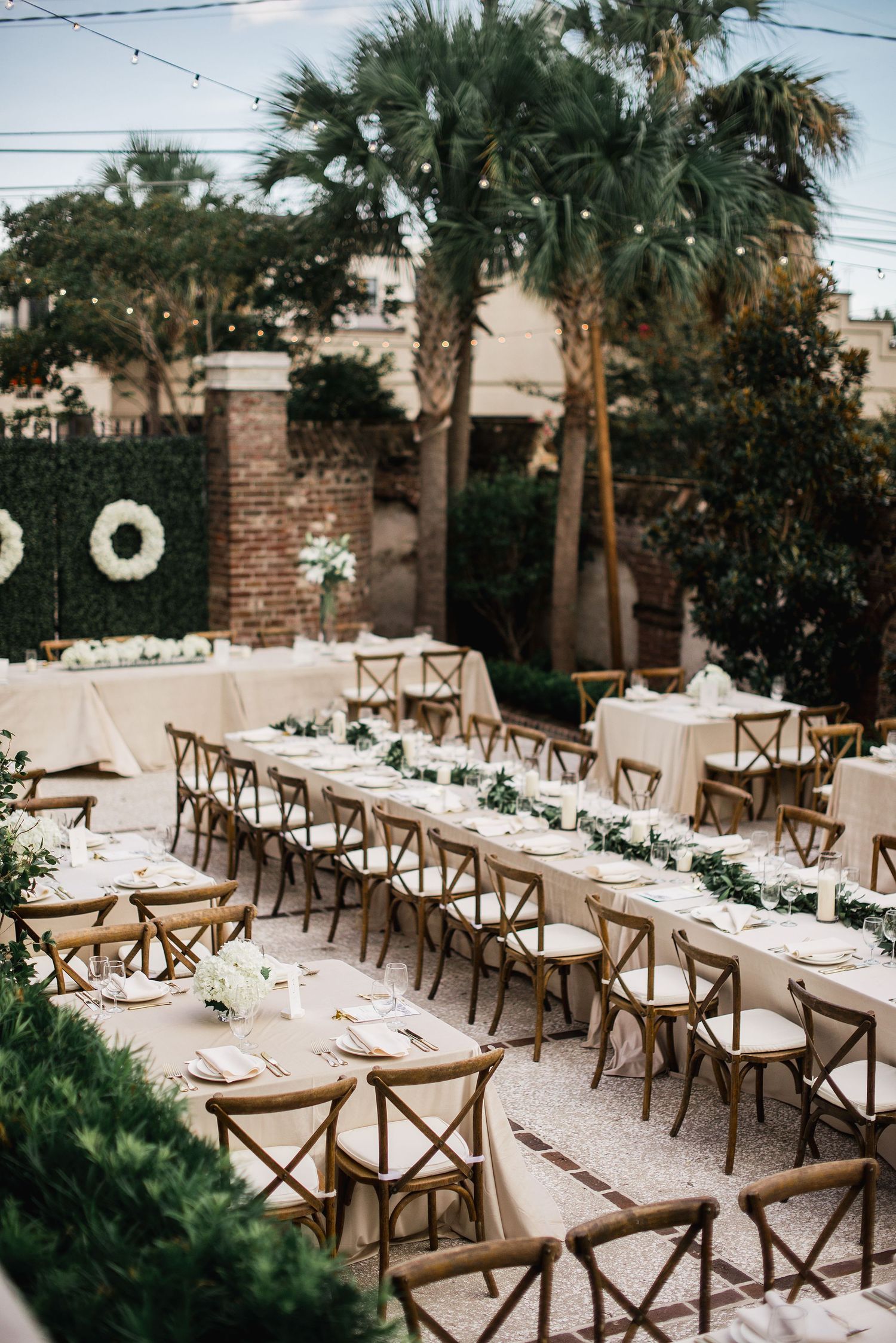 Long rustic wooden tables with white linens and greenery garlands set up for an elegant outdoor garden wedding reception.