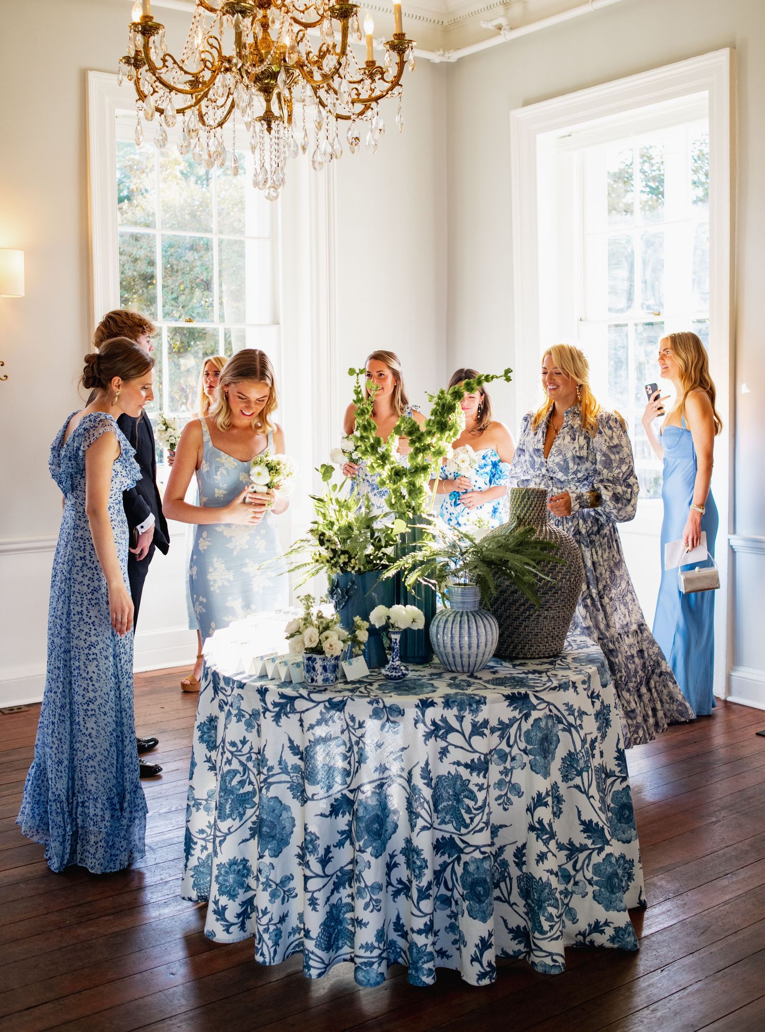 Wedding reception setup with elegant blue and white floral tablecloths and a crystal chandelier in a bright white room.