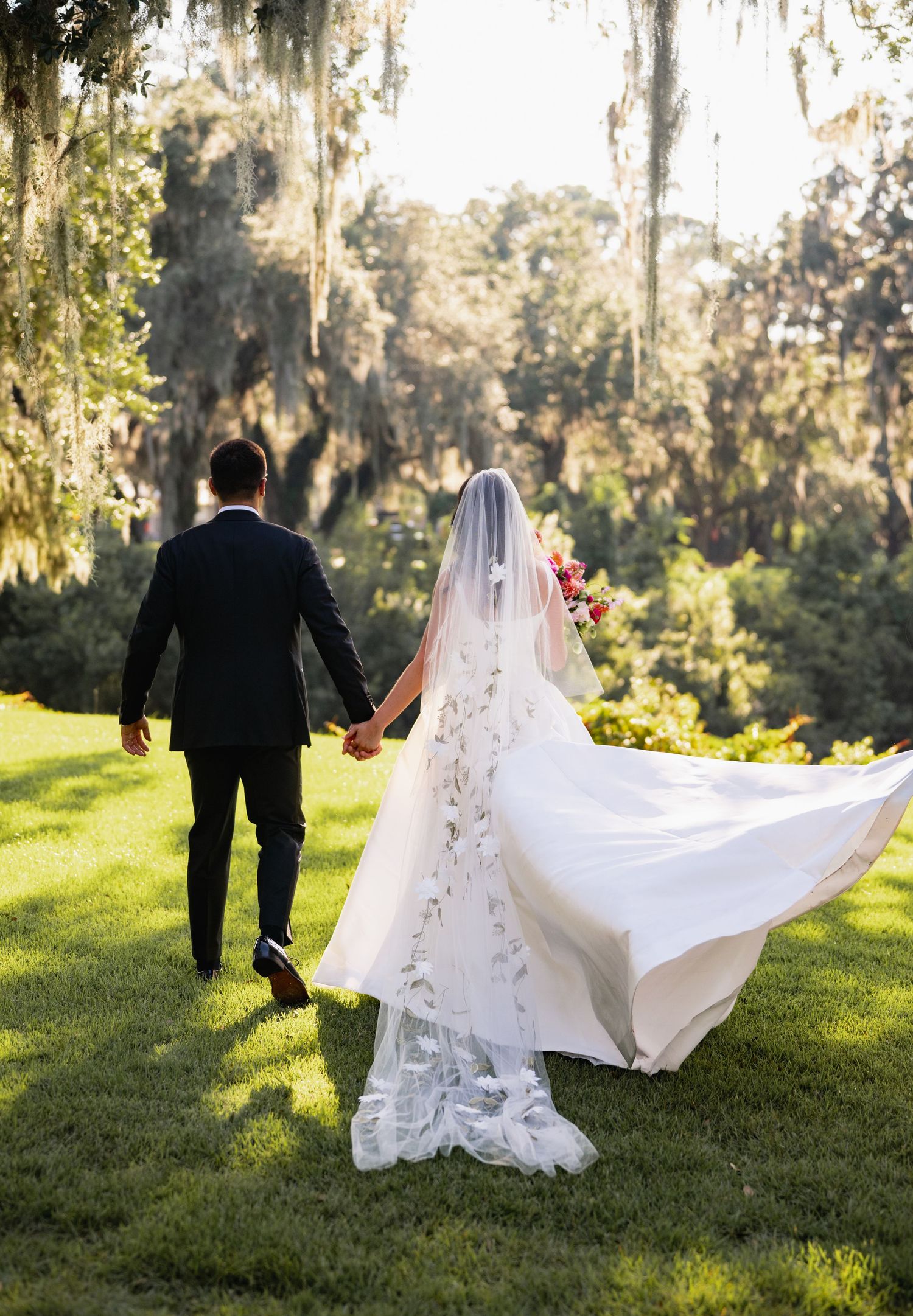 A couple in wedding attire walks hand in hand through a sunlit garden with Spanish moss draped trees in the background.