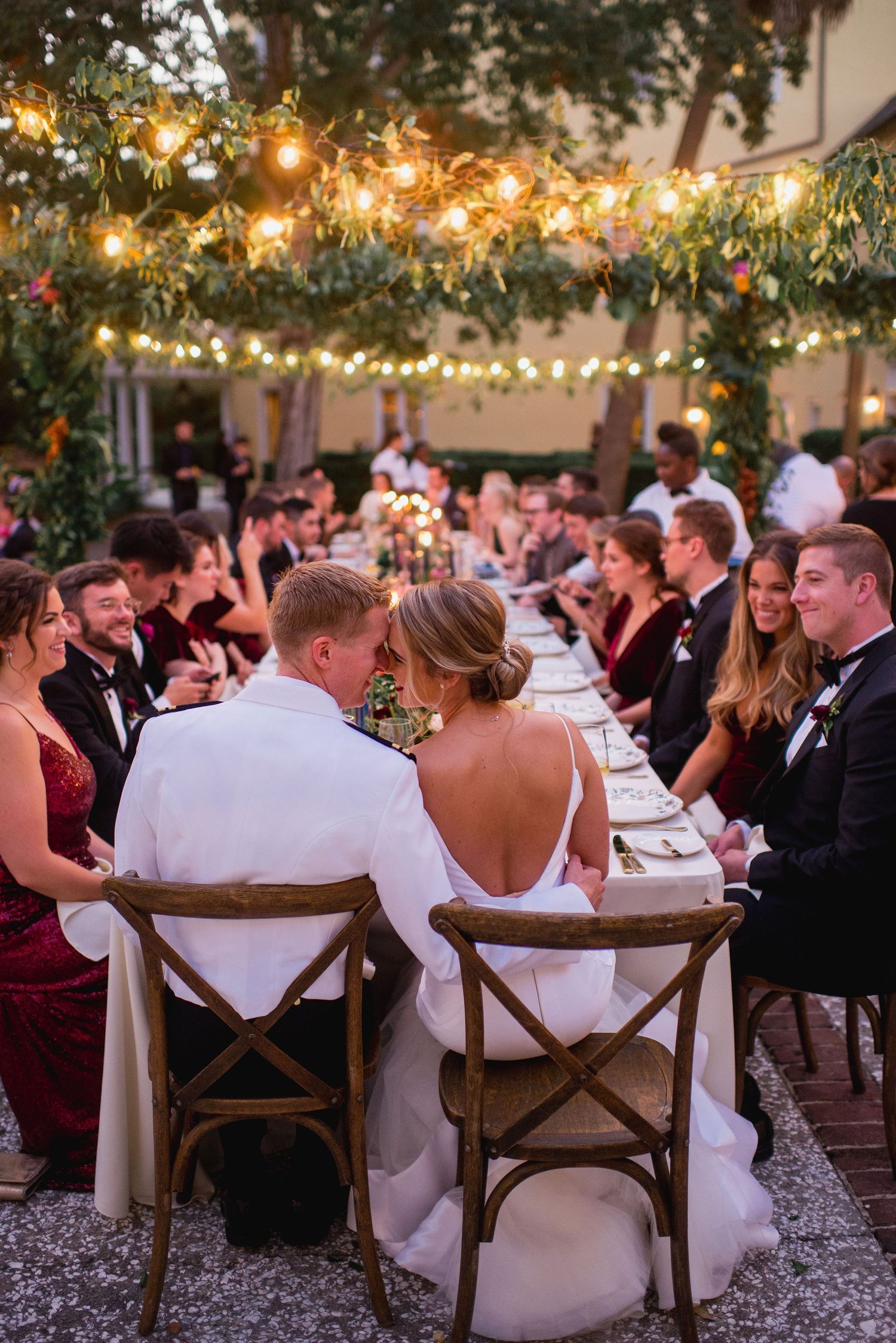 Guests seated at outdoor wedding reception tables under twinkling string lights at dusk.