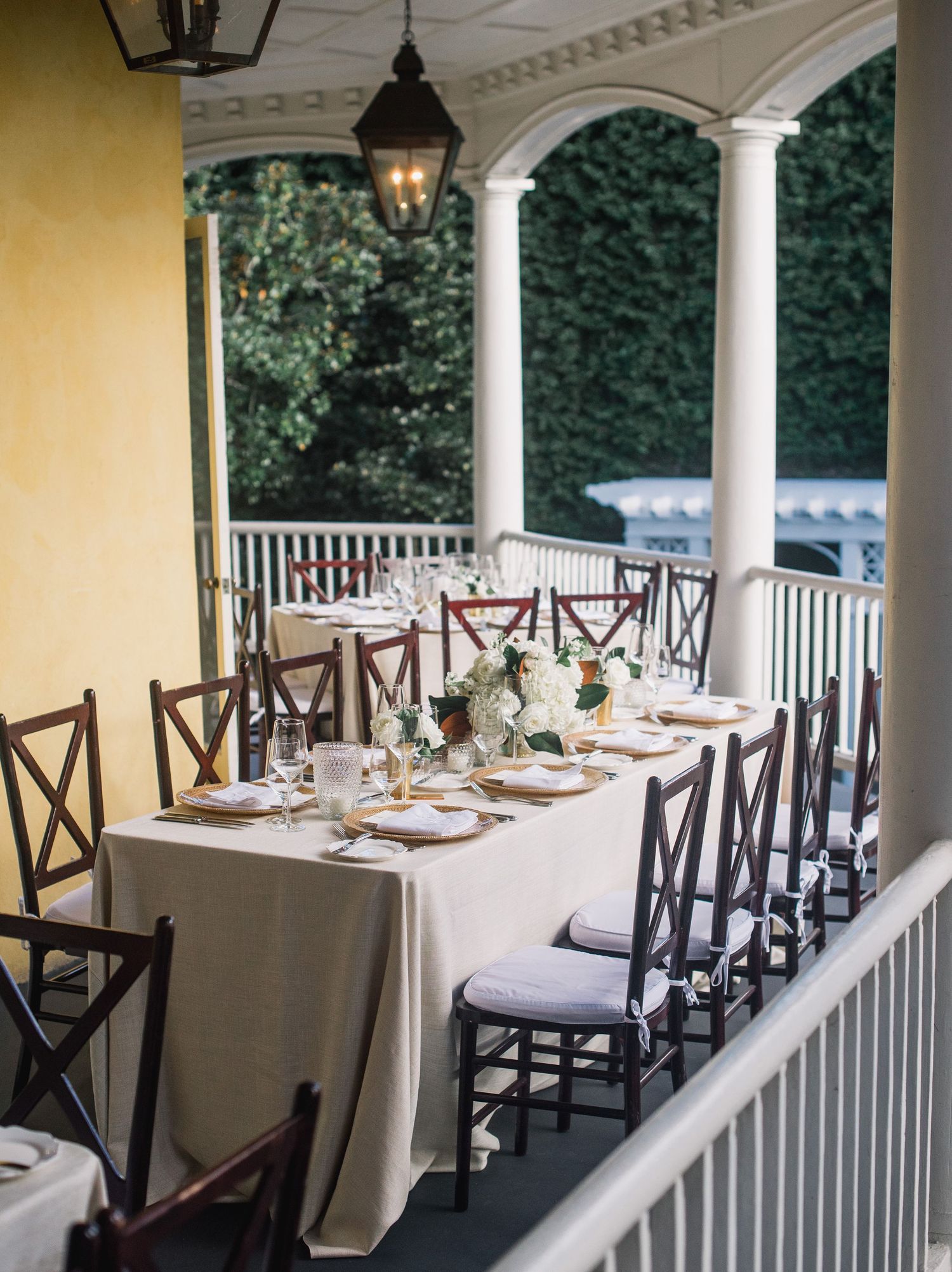 Elegant outdoor dining setup with white tablecloths and dark wooden chairs on a covered porch with columns and greenery.