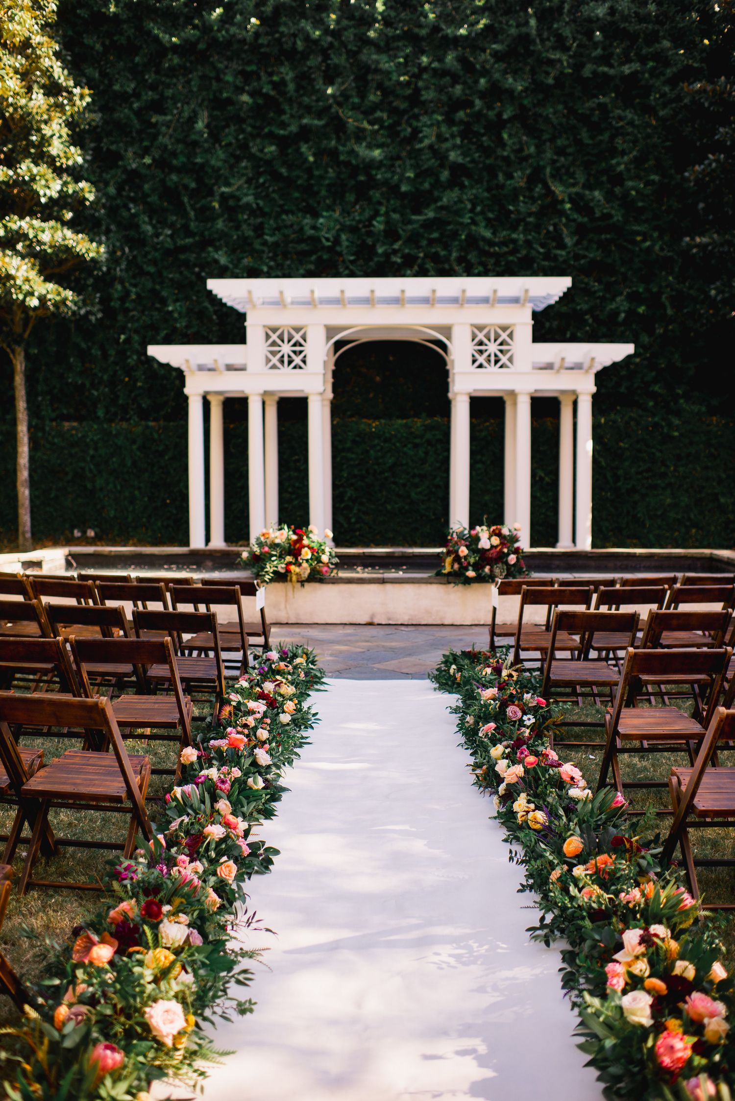 Elegant outdoor wedding ceremony setup with wooden chairs and colorful floral aisle arrangements leading to a white pergola.