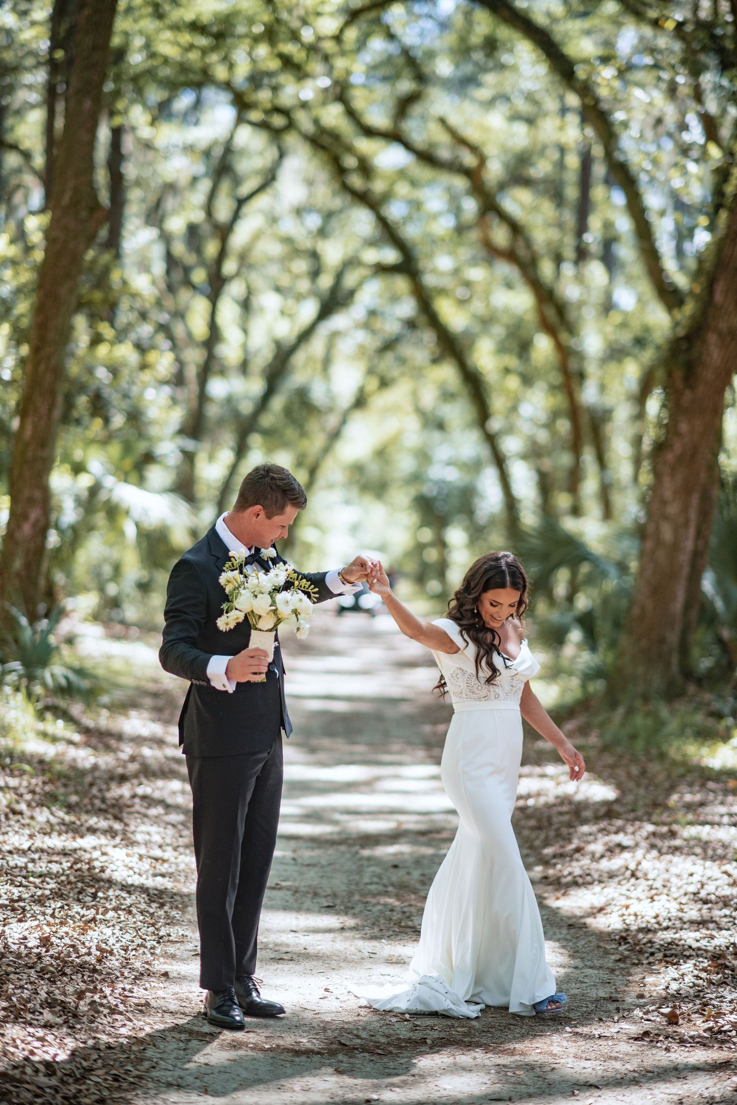A couple in wedding attire dancing together on a sunlit forest path surrounded by trees.