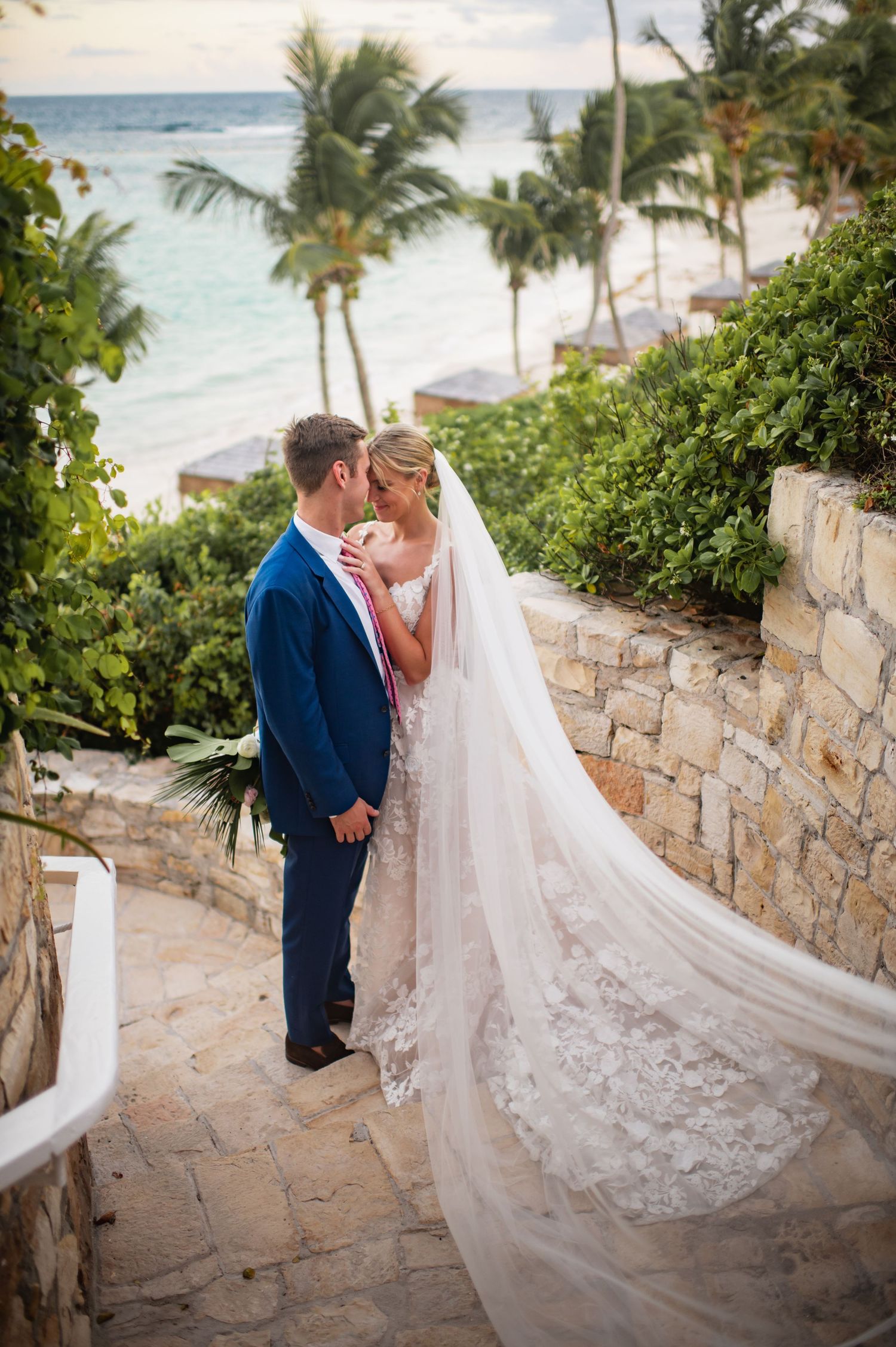 A romantic wedding couple walks down stone steps with ocean views and palm trees.