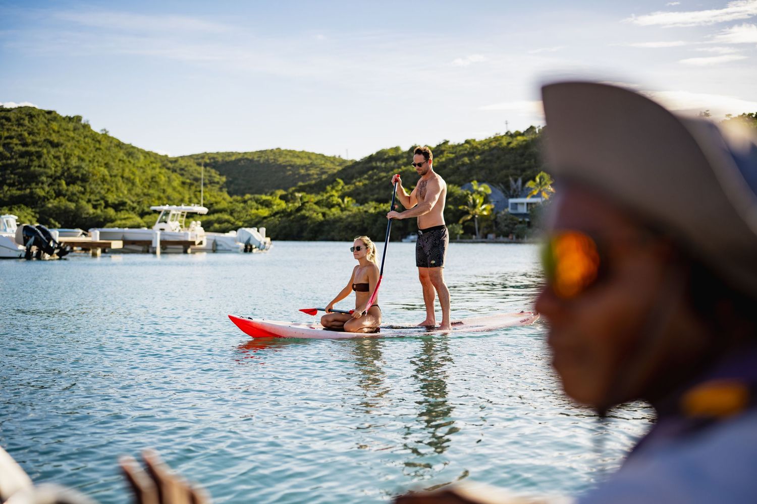 Vacationers stand on paddleboards in calm blue waters with green hills in the background.