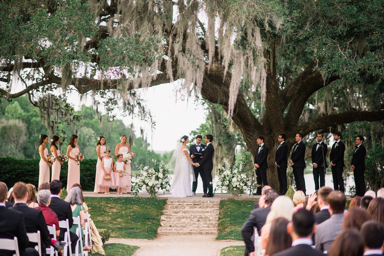 A wedding ceremony takes place under majestic oak trees draped with Spanish moss at an outdoor venue.