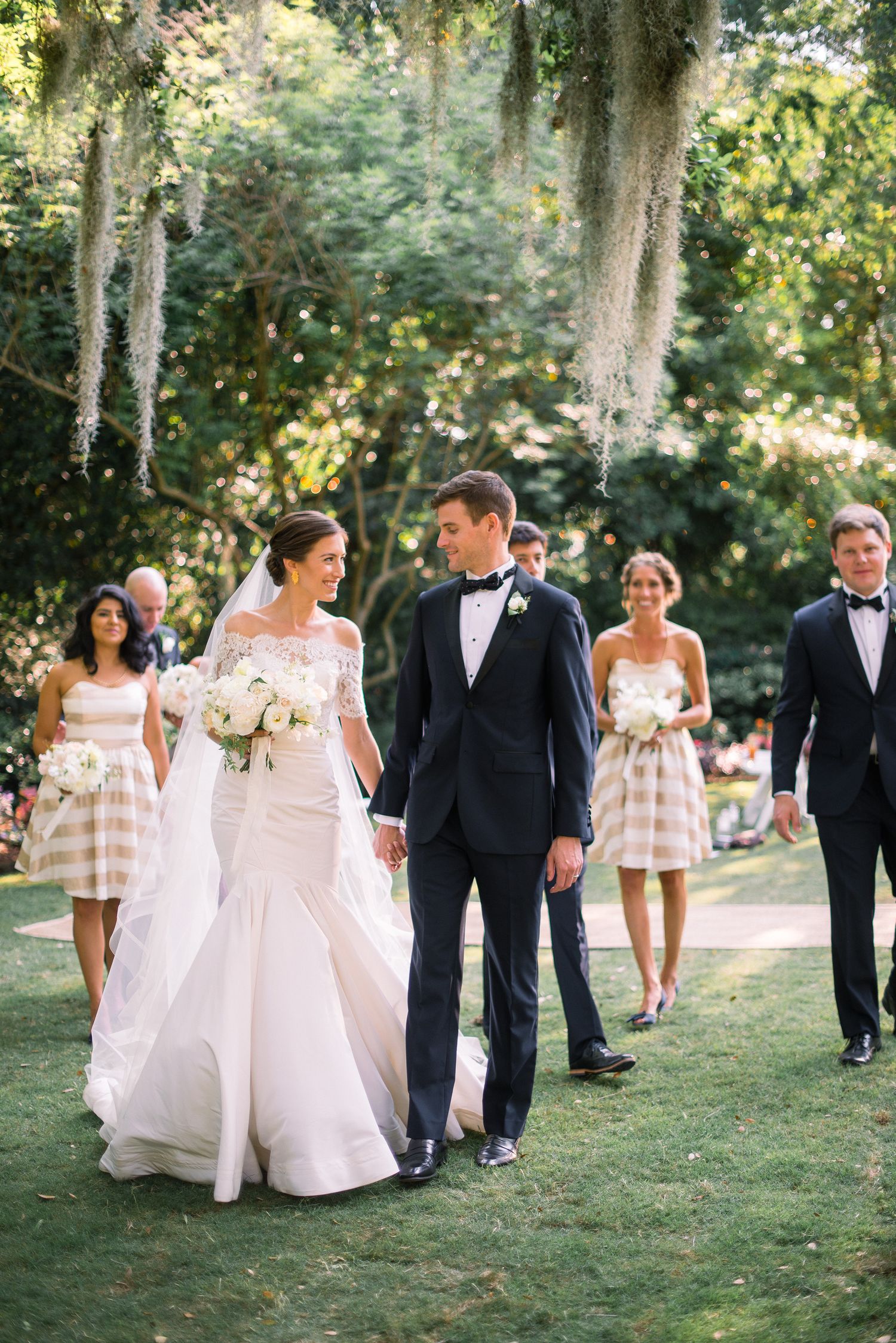 Wedding party walking on grass under Spanish moss draped trees in an elegant outdoor garden setting.