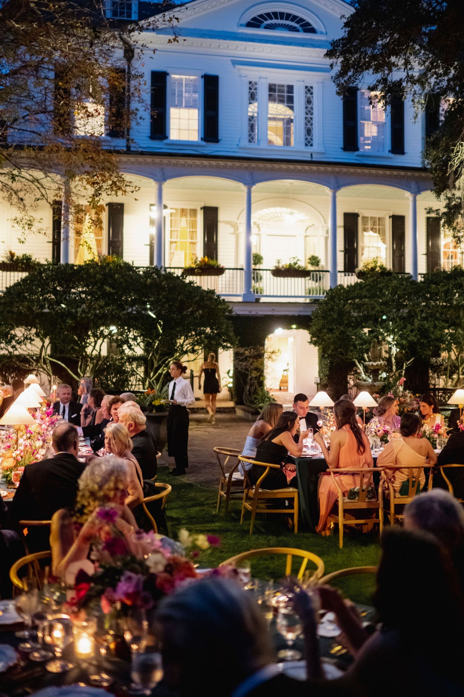 Evening outdoor wedding reception in a courtyard with string lights and guests dining under arched columns.