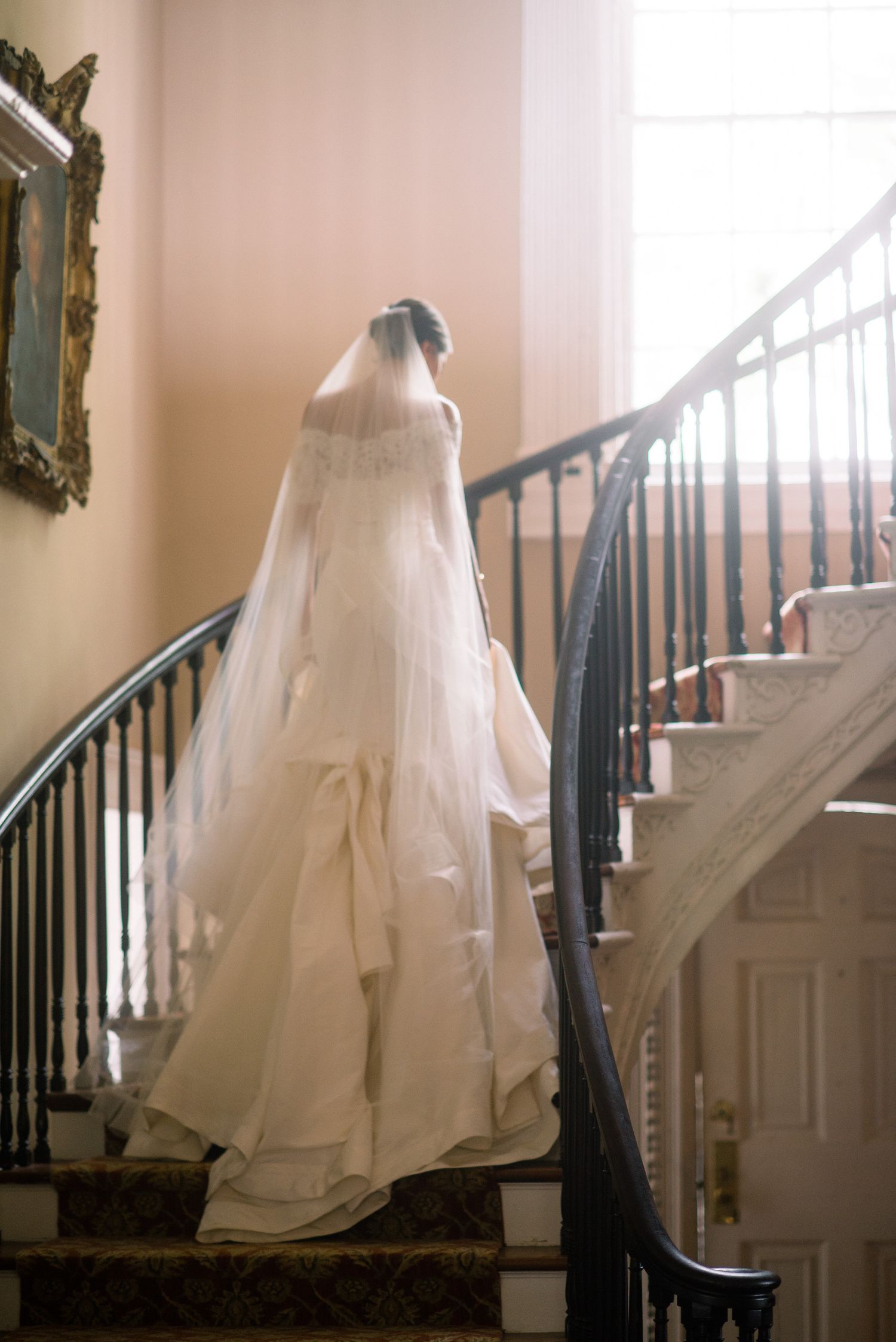 A flowing white wedding dress with ruffled layers cascades down an elegant staircase in soft natural light.