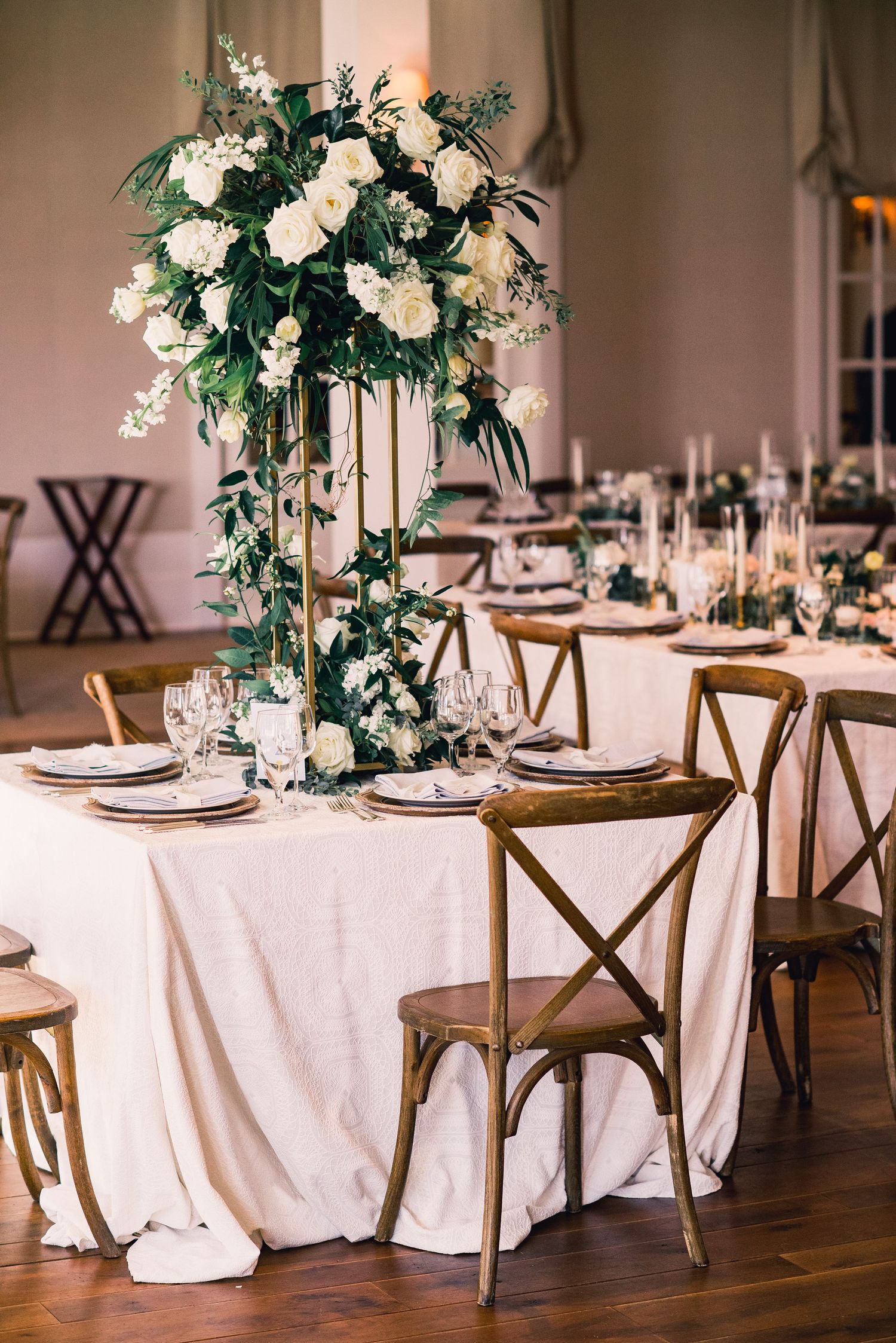 Elegant wedding reception tables with white linens, rustic wooden chairs, and tall white floral centerpieces in a ballroom setting.