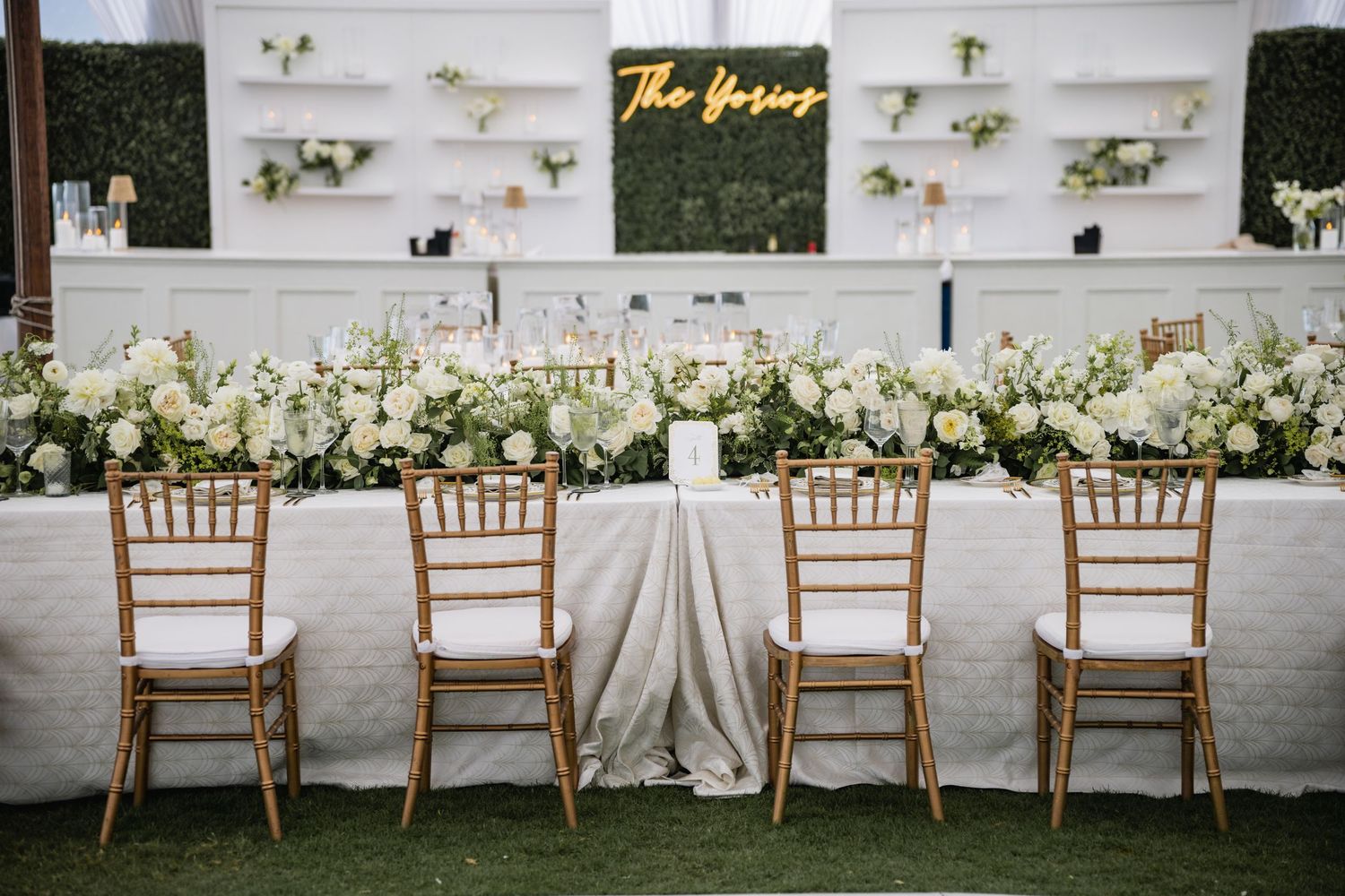 Elegant wedding reception table setup with wooden chairs and white floral arrangements against a decorative wall display.