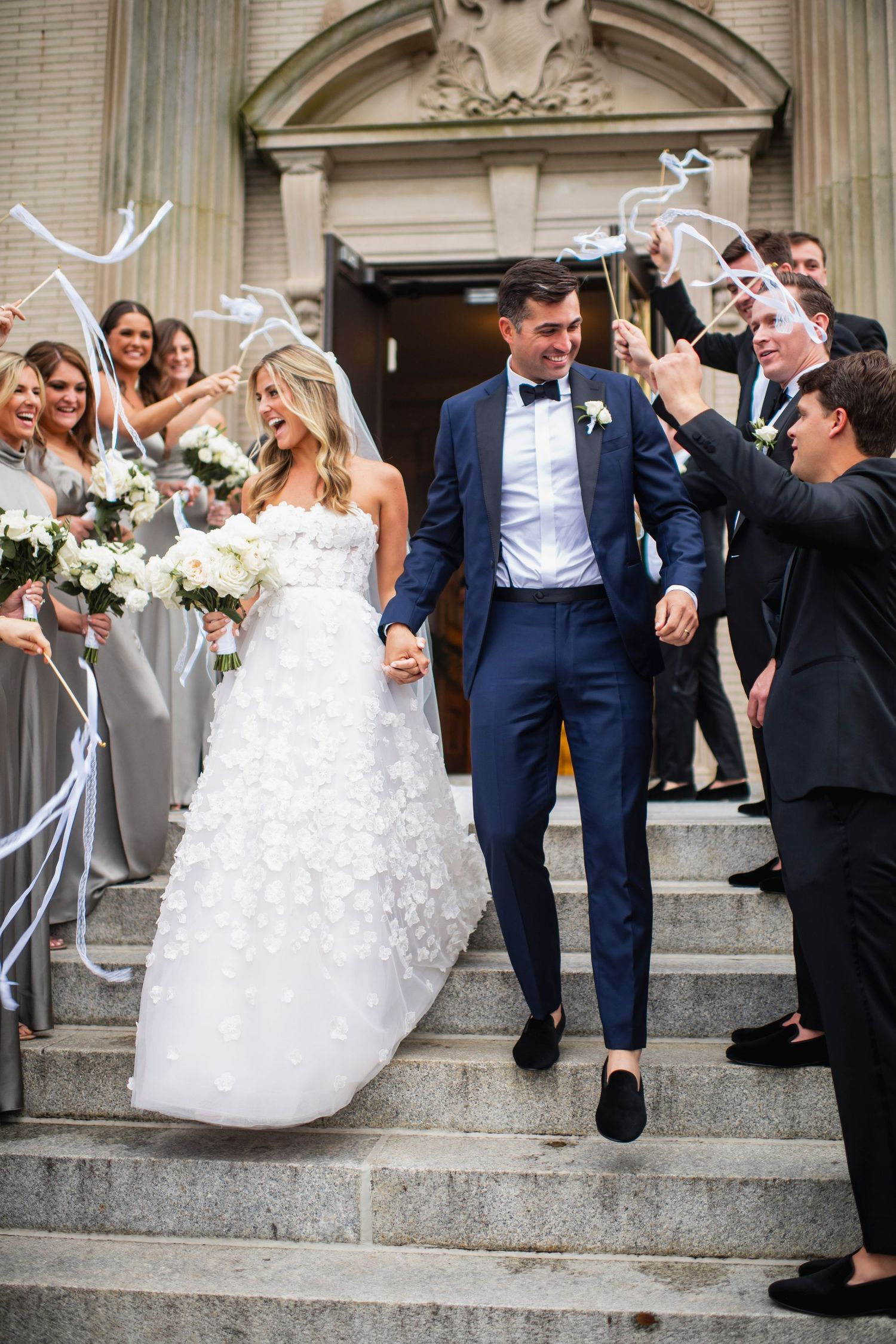 Wedding party descending church steps with the couple in white dress and navy suits.