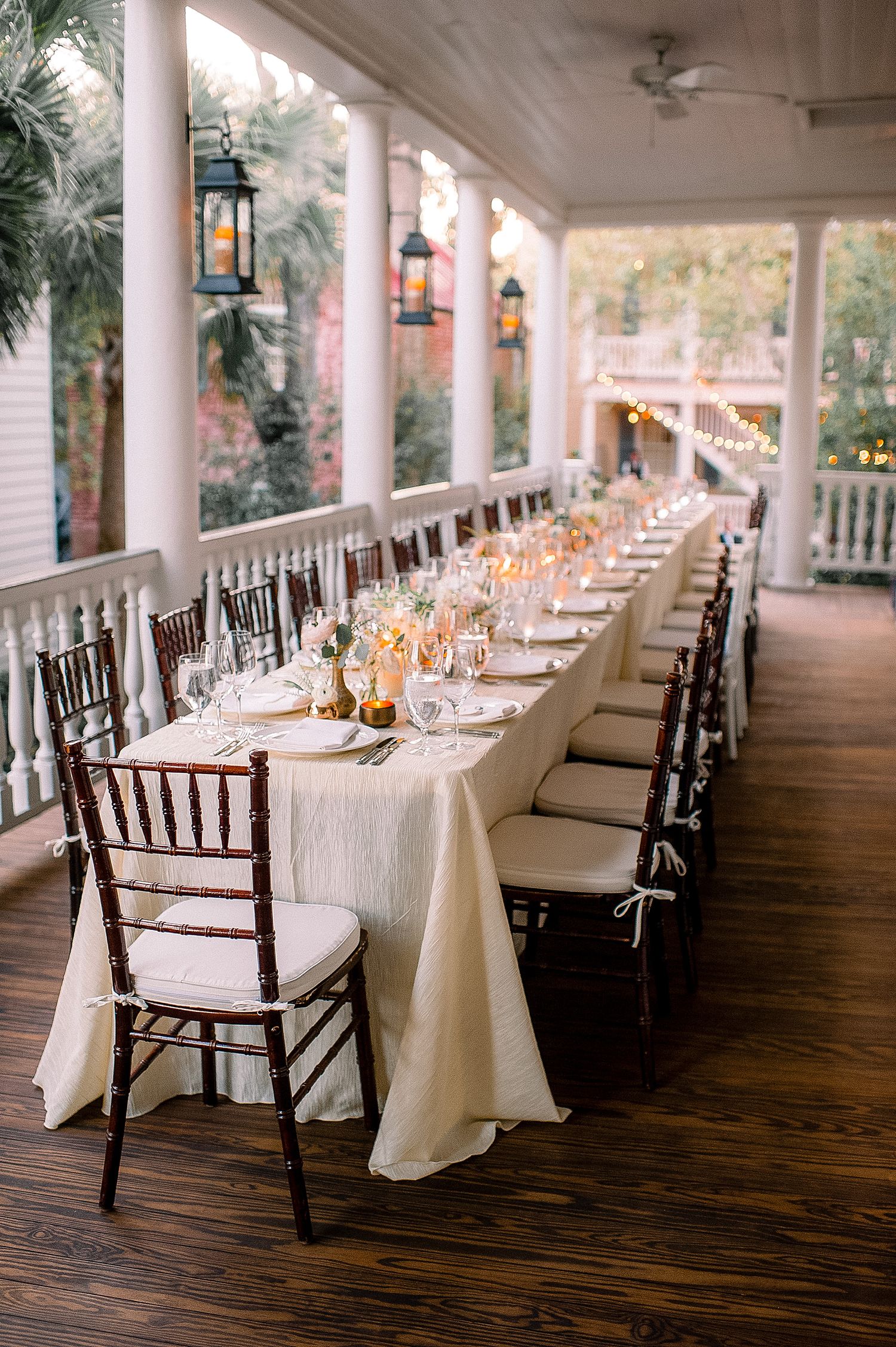 Long white dining tables with dark wooden chairs line an elegant covered porch for an outdoor wedding reception setup.