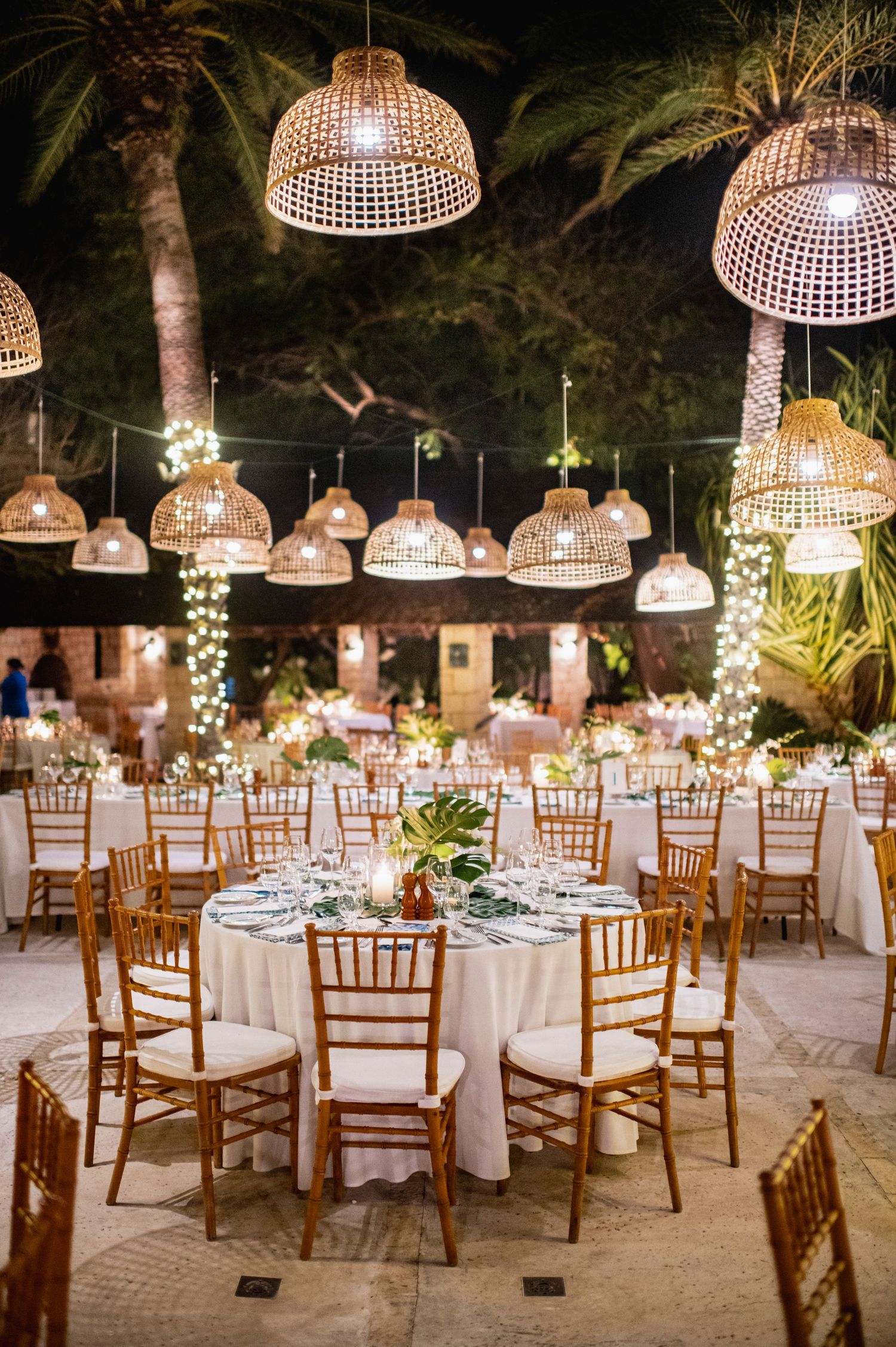 Romantic outdoor wedding reception venue with wicker lanterns and wooden chairs under palm trees at night.