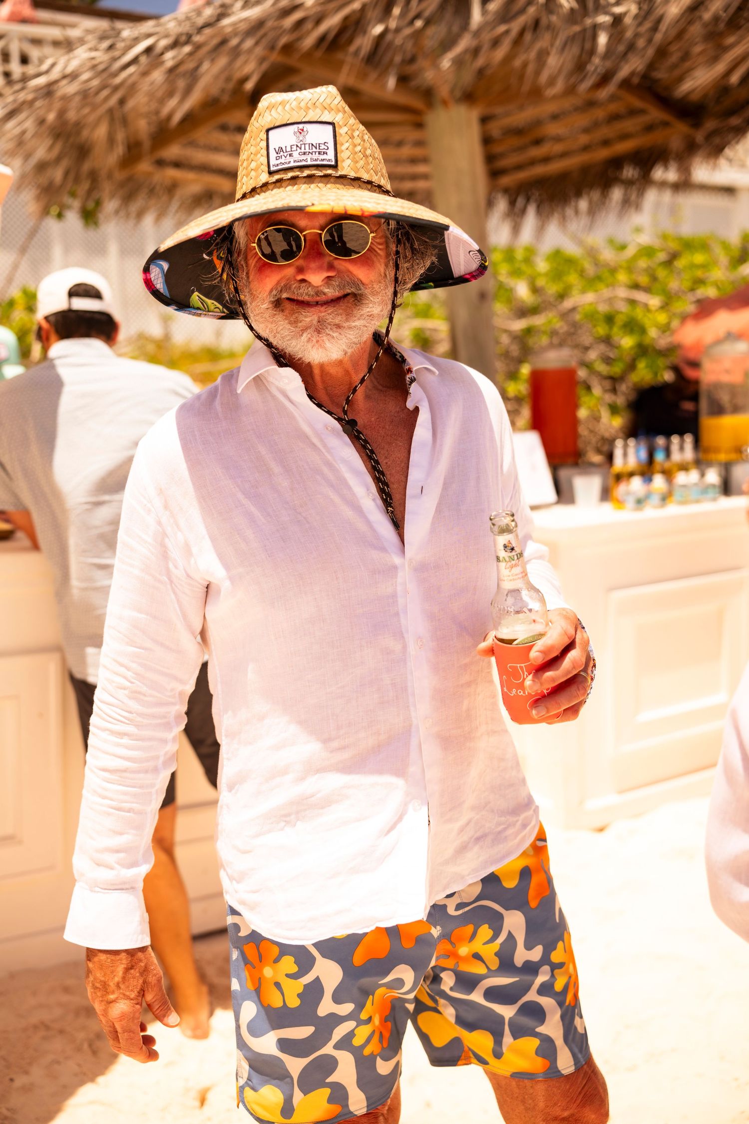 Beach party guest in casual attire wearing a straw hat and sunglasses under a thatched umbrella.