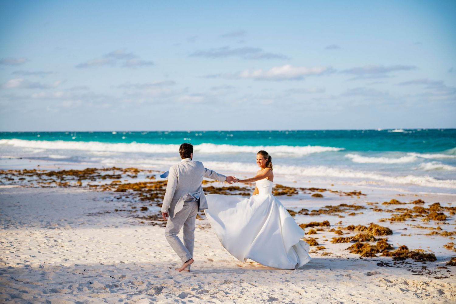 Newlyweds walking hand in hand on beach with white dress flowing in ocean breeze.