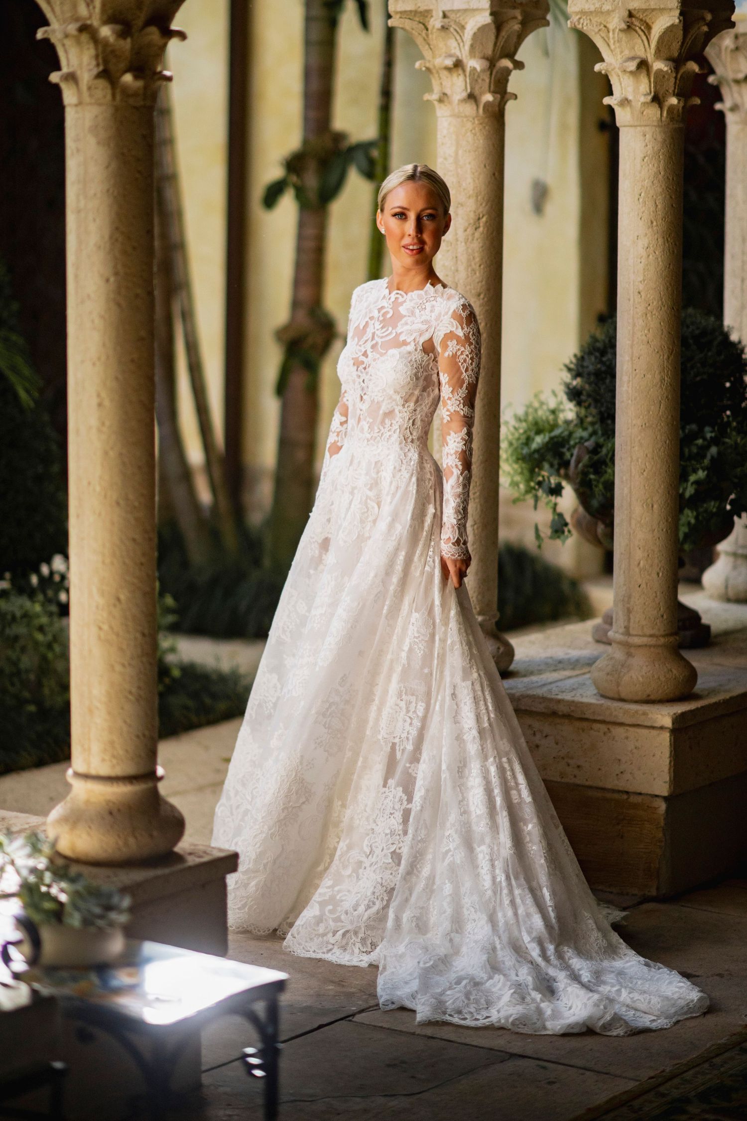 Elegant lace wedding dress with long train photographed in a columned courtyard setting.