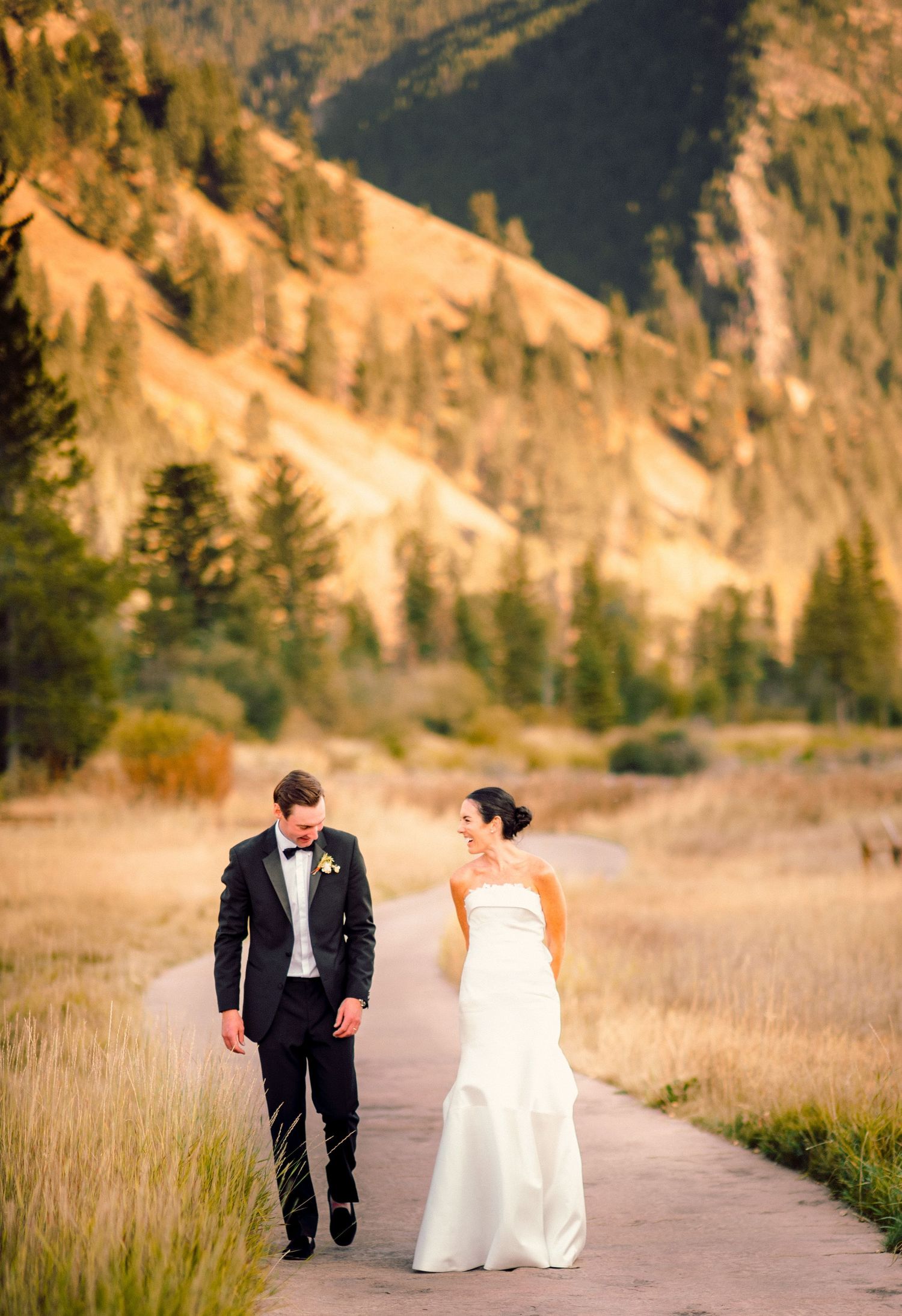 A couple in wedding attire walks along a scenic mountain path during golden hour in a stunning outdoor setting.