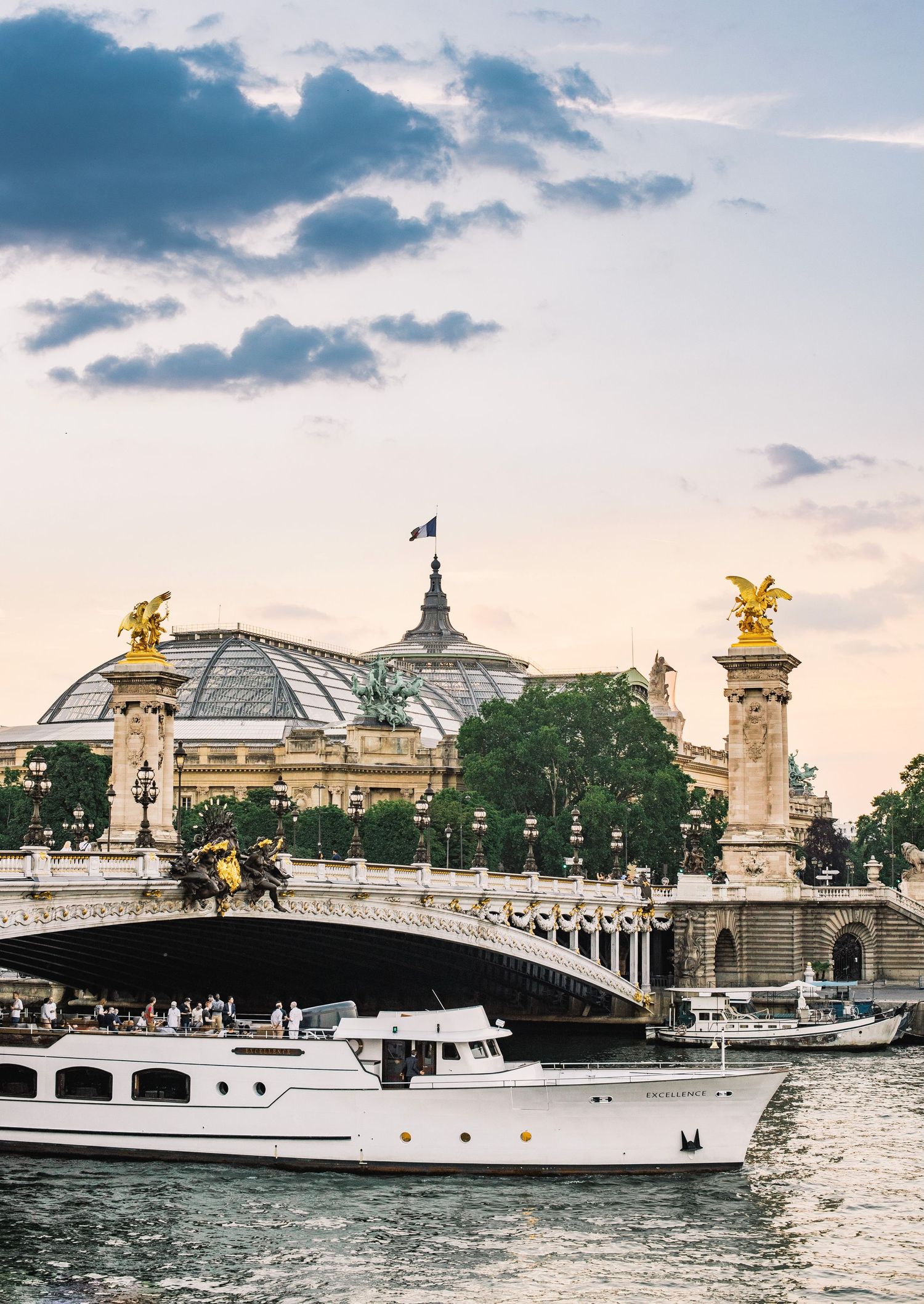 Luxury yacht passing under the Pont Alexandre III bridge near the Grand Palais in Paris at sunset.