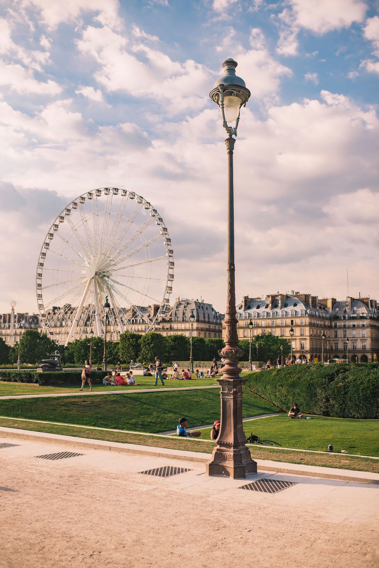 Ferris wheel and ornate lamp posts stand against a cloudy sky in a scenic Paris park setting.
