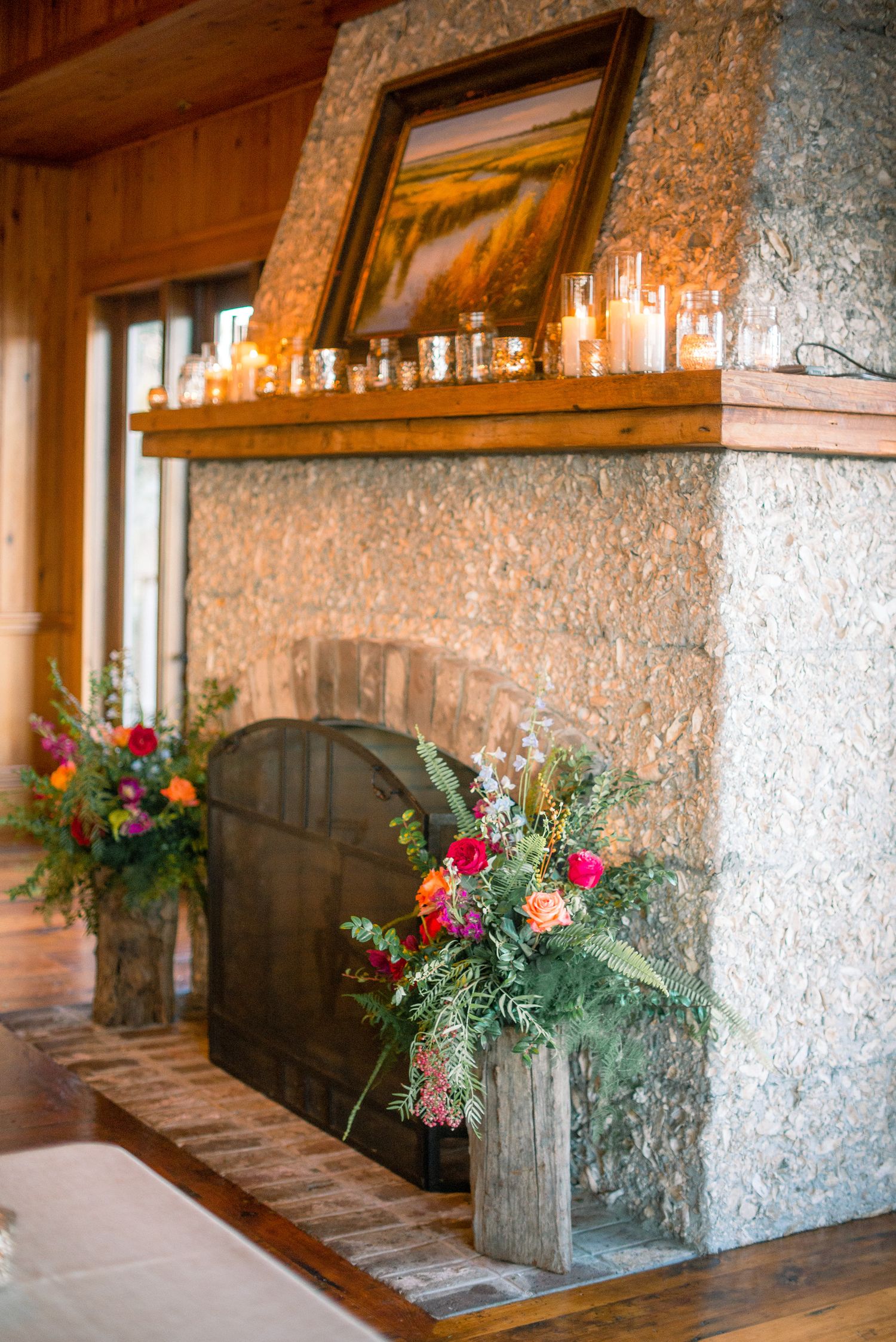 Rustic stone fireplace with wooden mantel decorated with candles and colorful floral arrangements in wooden boxes.