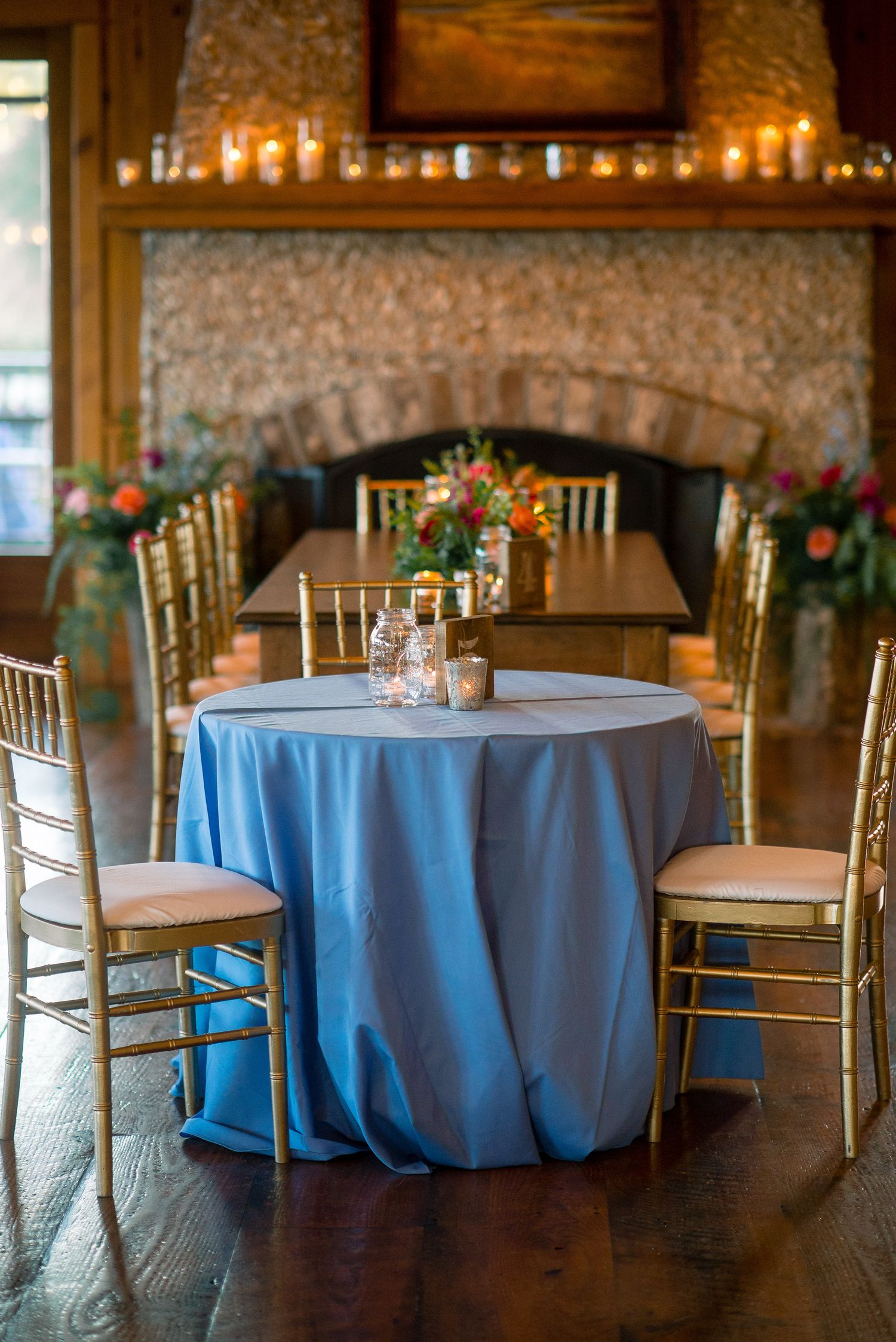 Round tables with light blue tablecloths and gold chiavari chairs set up in an elegant rustic venue with stone walls.