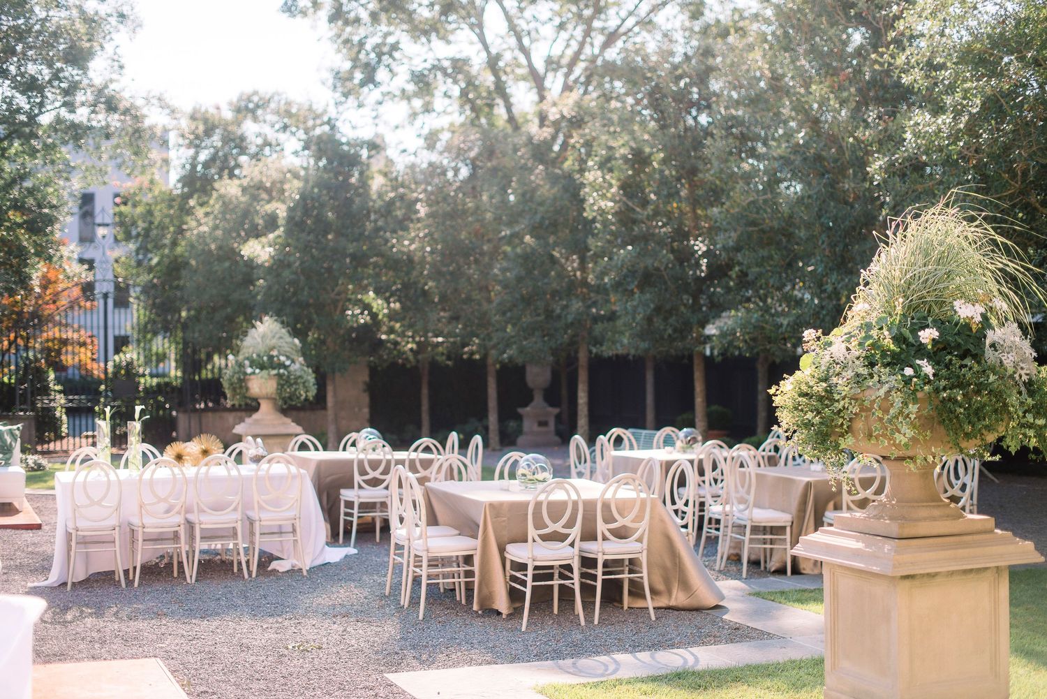 Elegant outdoor wedding reception setup with white chairs and tables arranged under natural tree canopy lighting.