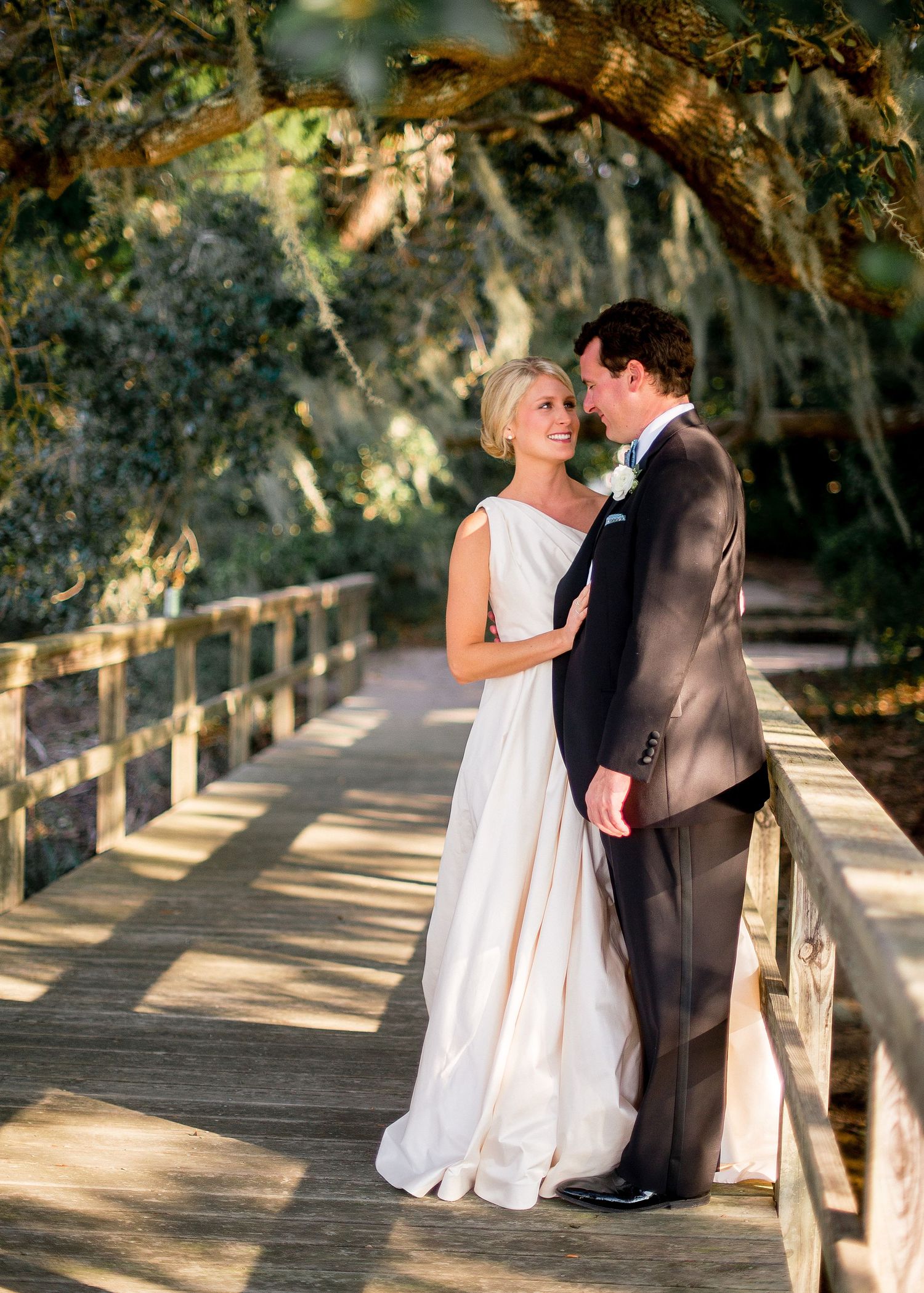 A romantic couple shares an intimate moment on a sunlit wooden boardwalk surrounded by lush greenery.