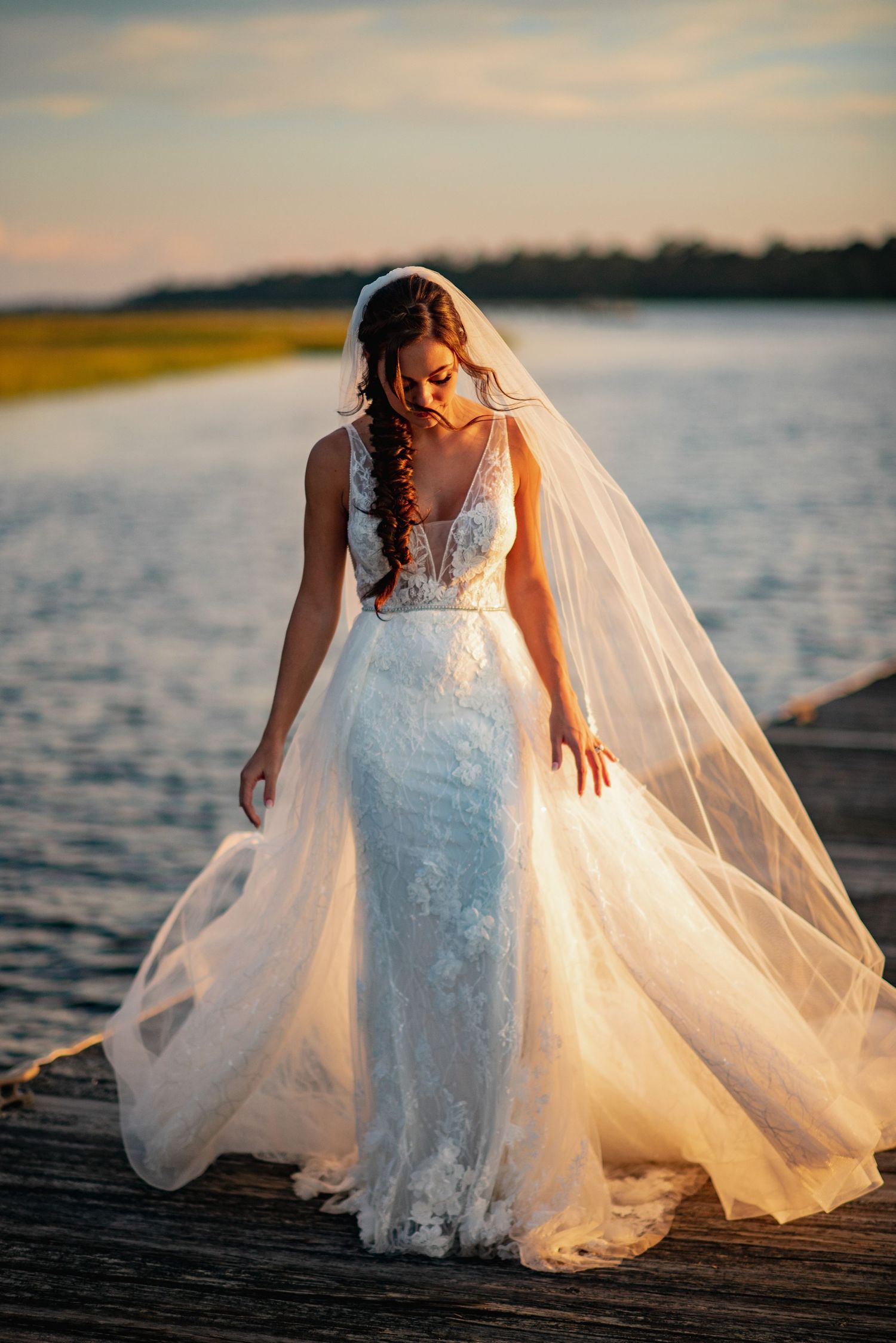 A bride in a lace wedding dress and flowing veil stands on a dock at sunset with water and golden light behind her.